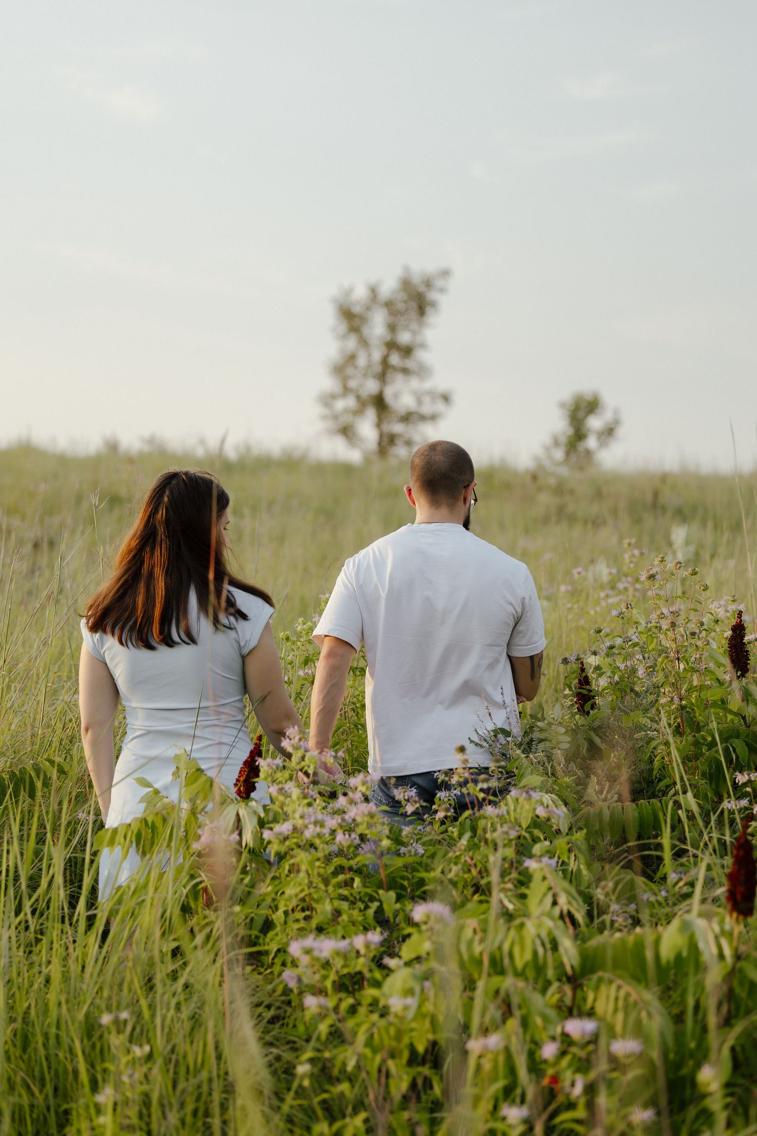 Couple in white outfits walking hand in hand through a lush green wildflower meadow on a sunny day.