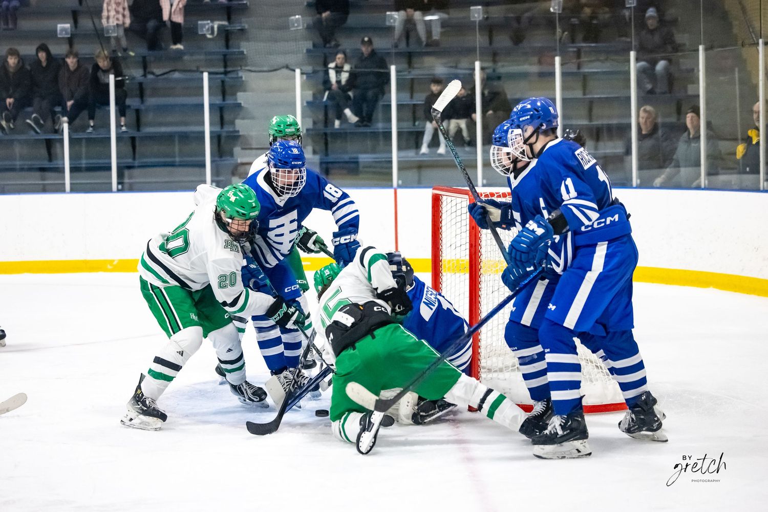 Youth hockey players in blue and green uniforms battle for puck near goal.