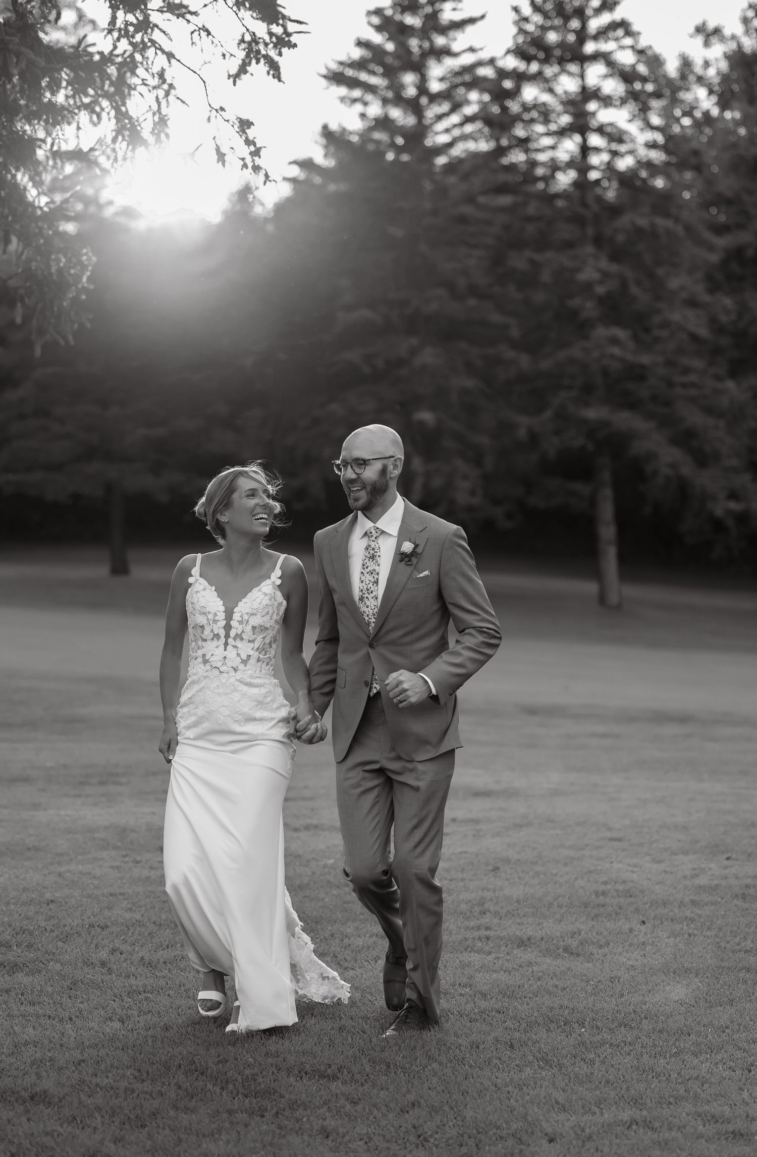 Black and white photo of laughing bride and groom walking hand in hand across golf course at sunset.