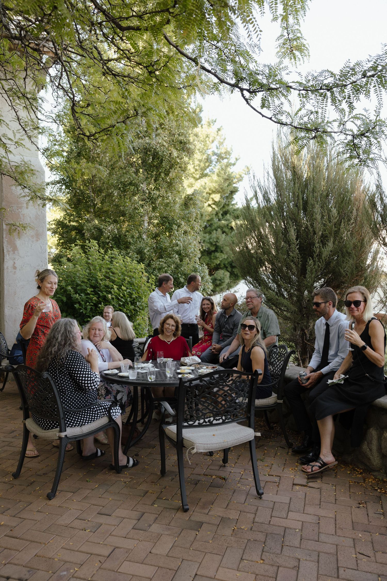 Wedding guests socializing outdoors at patio tables surrounded by lush greenery.