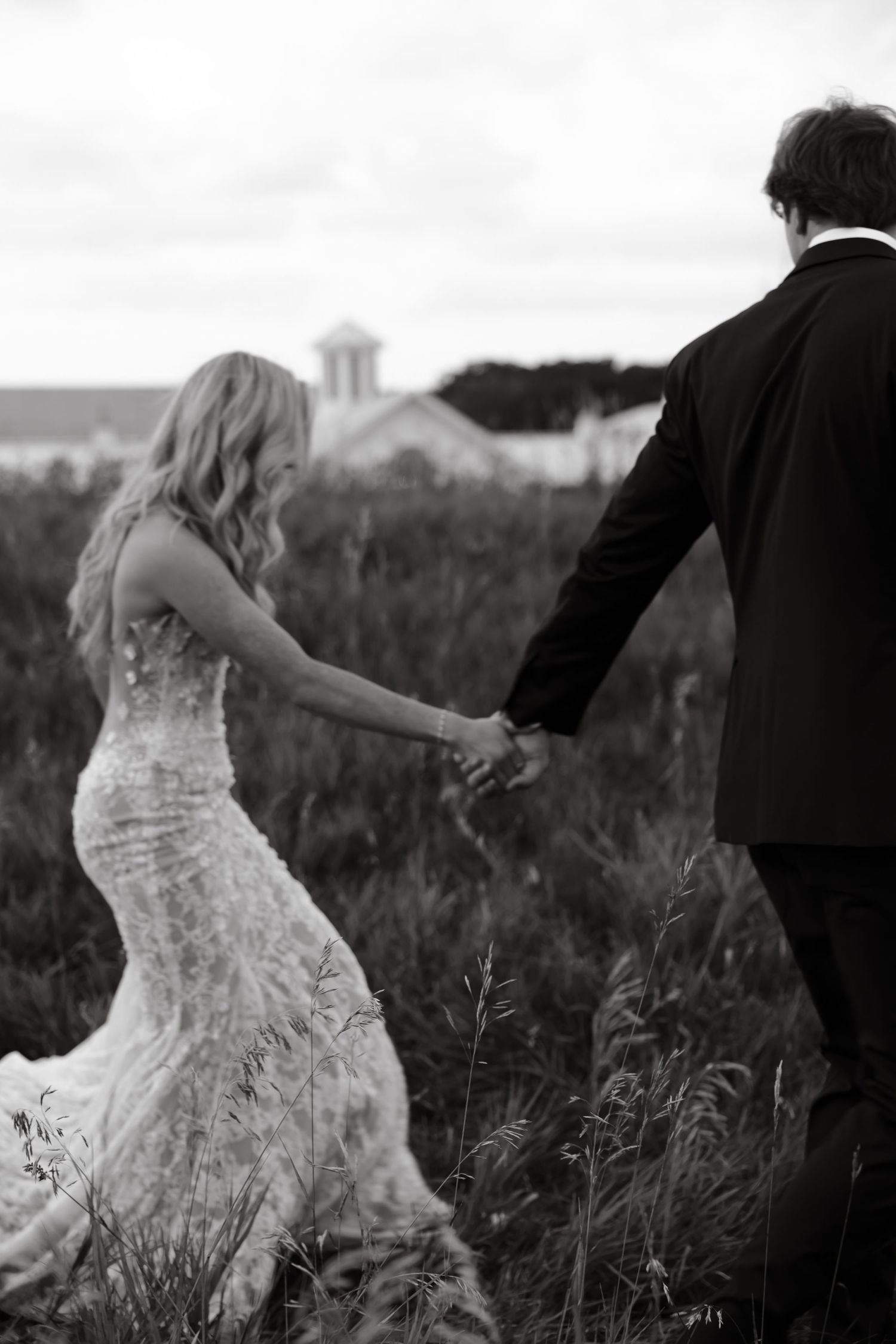 Black and white photo of bride and groom holding hands walking through tall grass field.