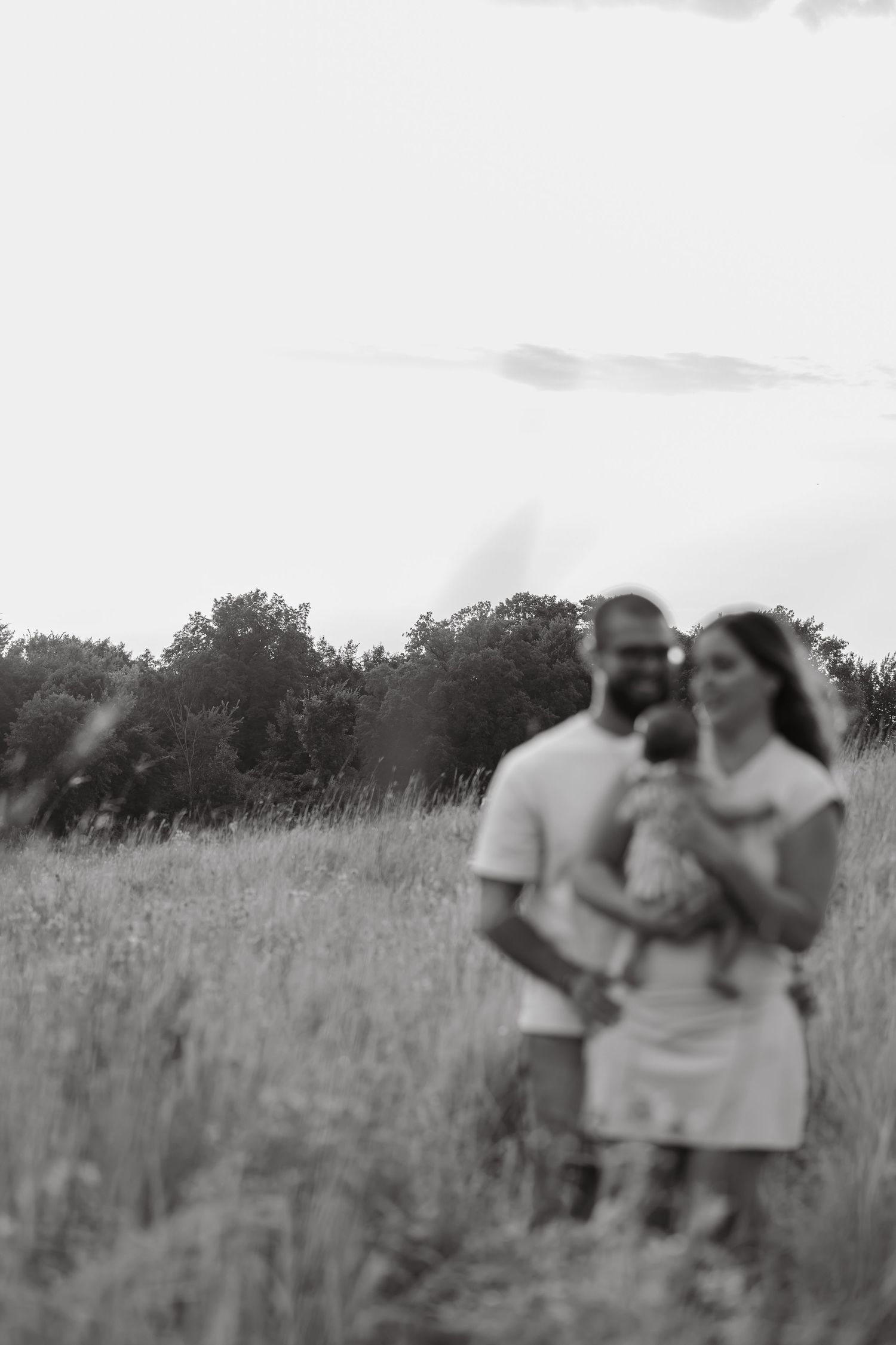 Black and white photo of a couple holding a baby in a field with trees in the background.