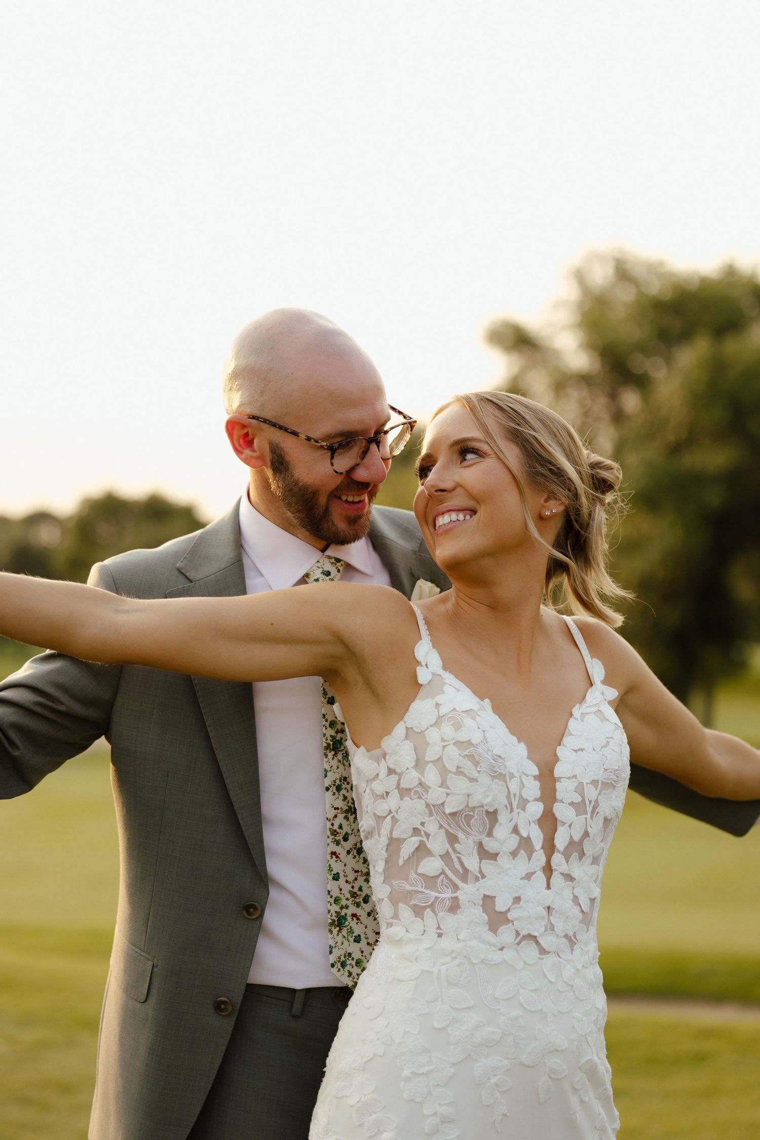 Smiling bride in lace gown embraces bald groom in gray suit with floral tie at golden hour outdoor wedding.