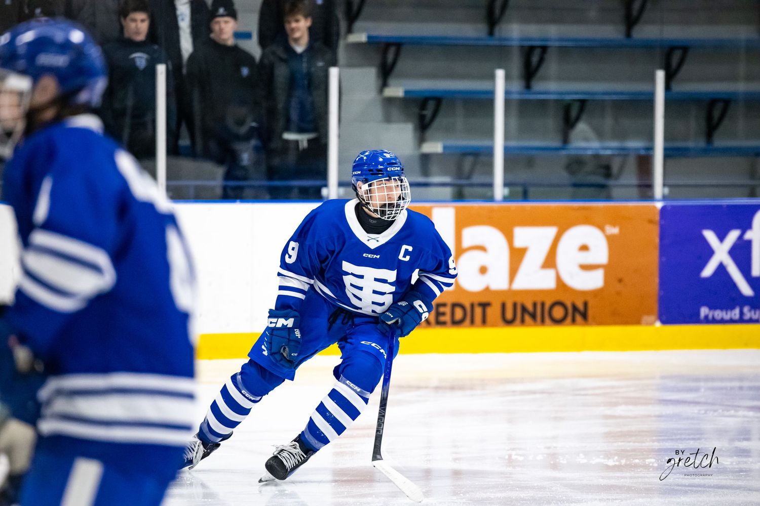 Youth hockey team captain in blue uniform skating on indoor ice rink.