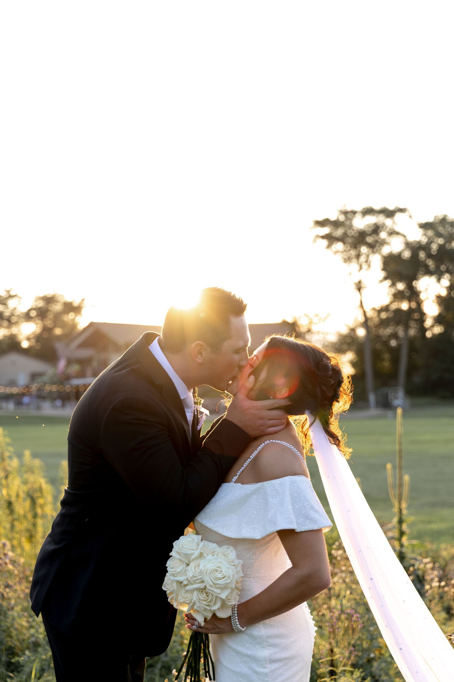 Bride and groom kissing at golden hour sunset, bride in off-shoulder gown holding white bouquet.