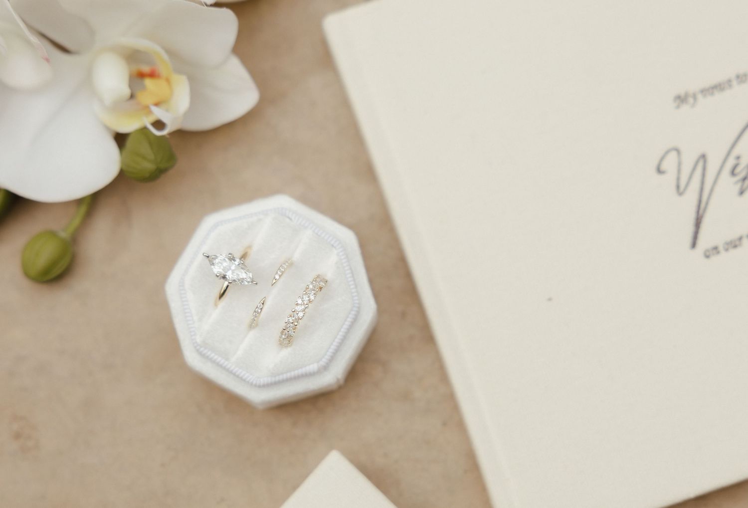Elegant wedding rings displayed in a white velvet ring box beside vow booklet and white orchid.