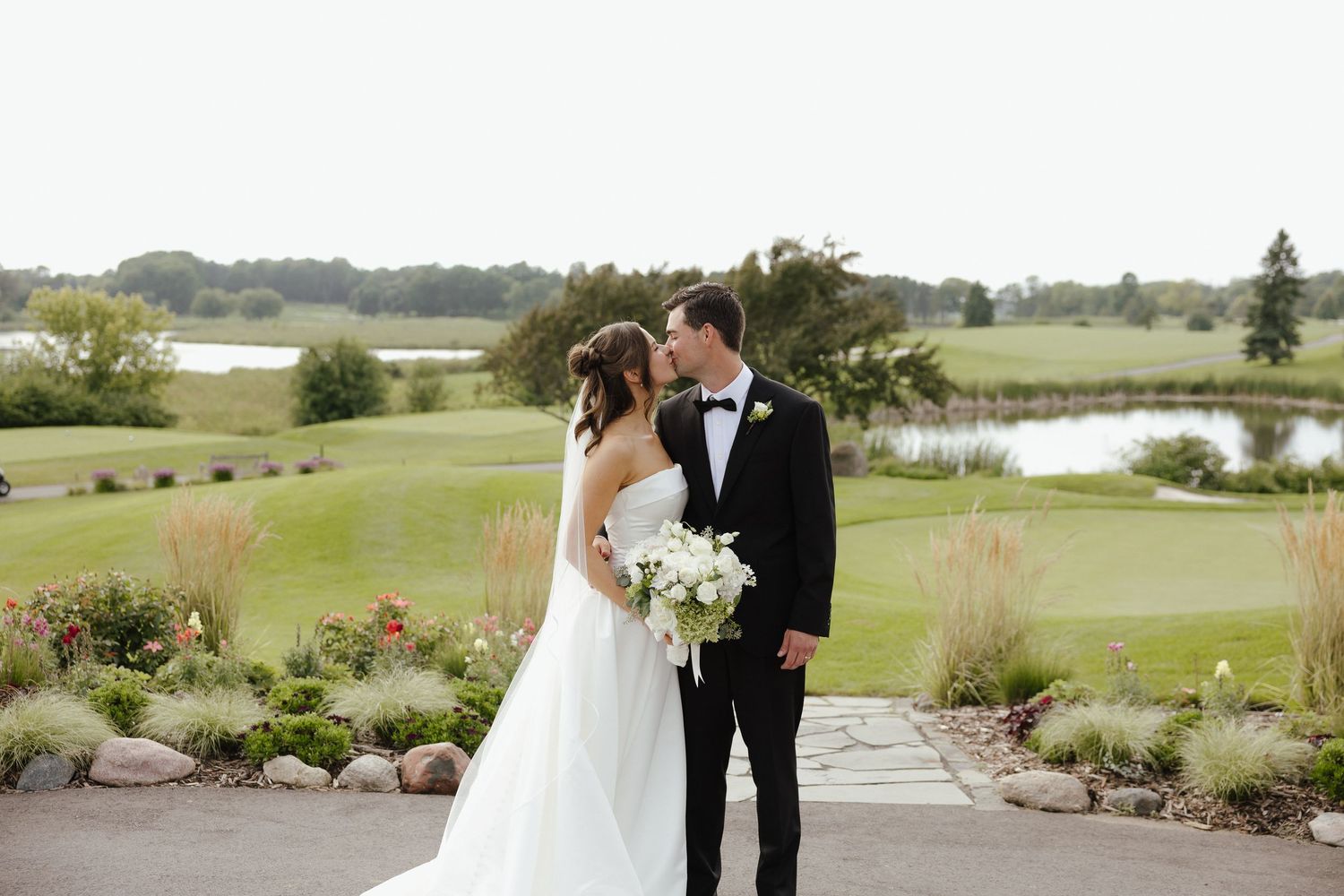 Newlywed couple sharing a kiss overlooking a scenic golf course with pond and lush greenery on their wedding day.