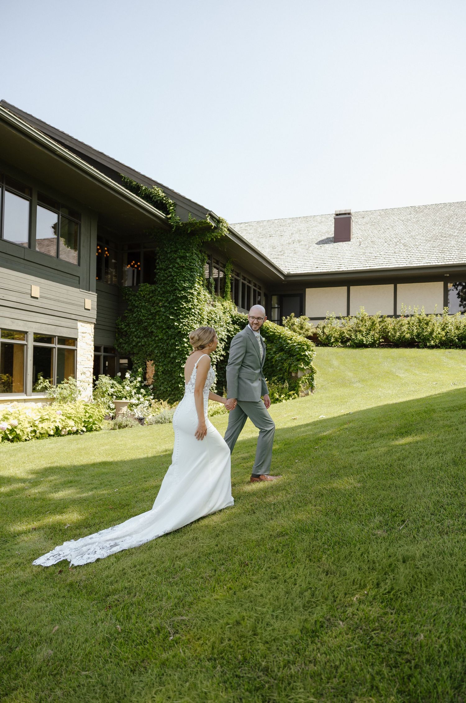 Bride and groom walk hand in hand across lush green lawn outside ivy-covered wedding venue on sunny day.