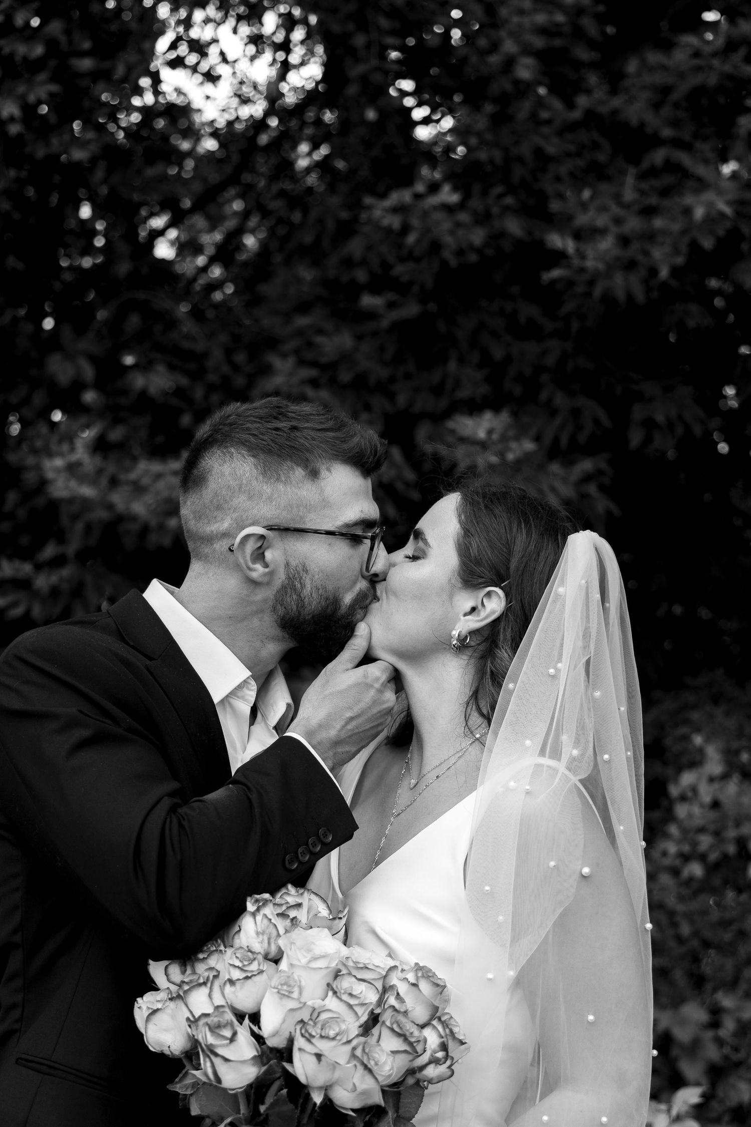 Black and white photo of groom kissing bride holding rose bouquet in garden setting.