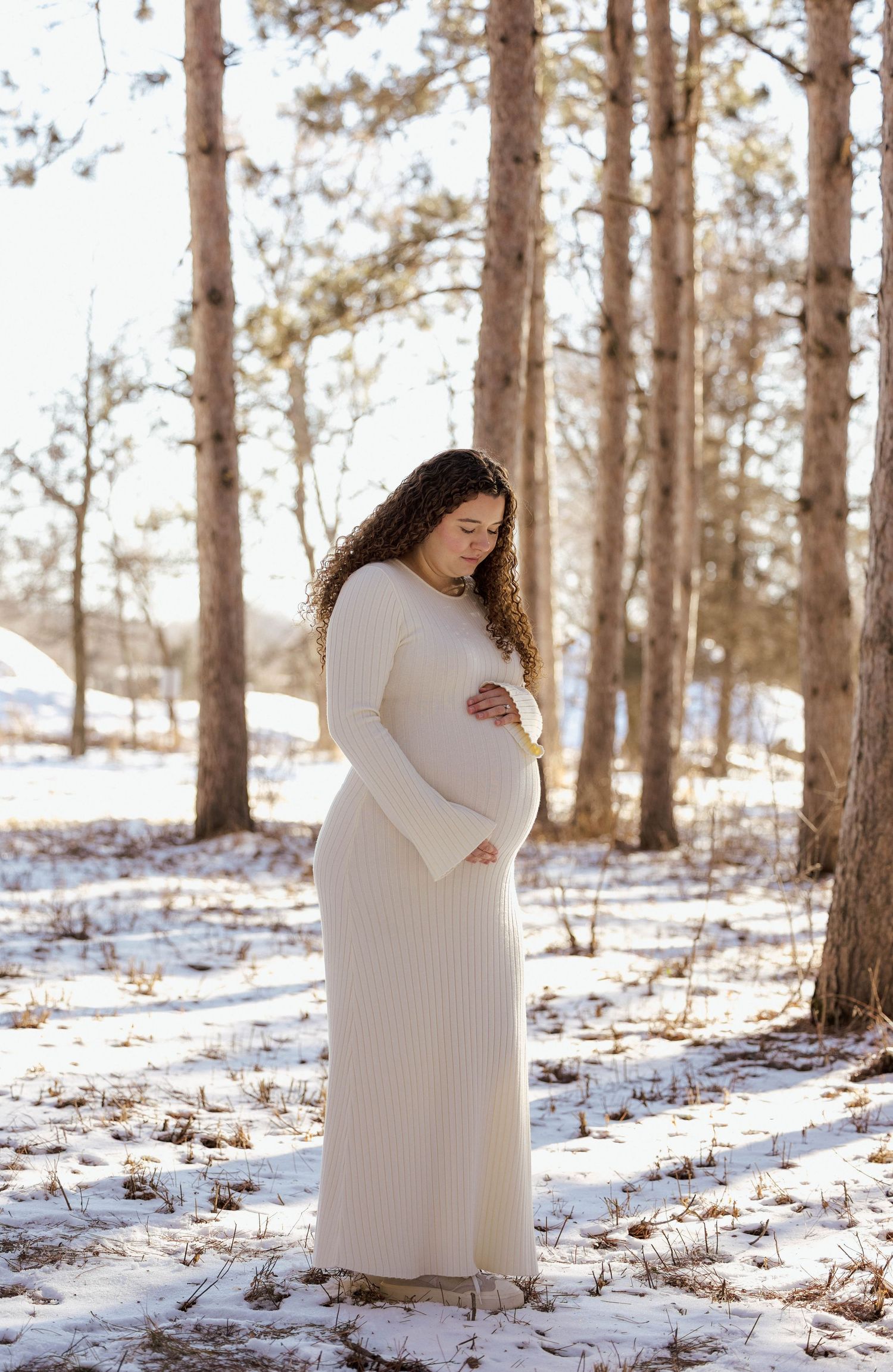 Pregnant woman in cream ribbed maxi dress standing among trees in snow.