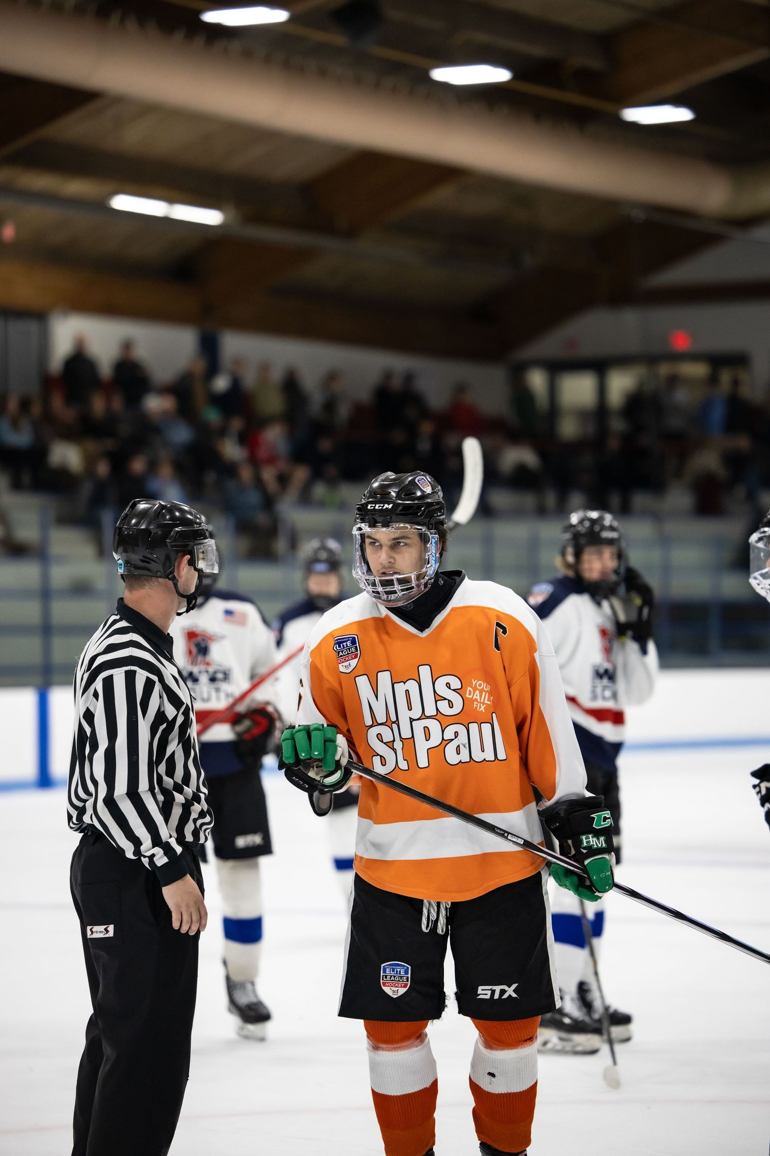 Hockey player in orange Mpls St. Paul jersey speaking with referee on ice rink.