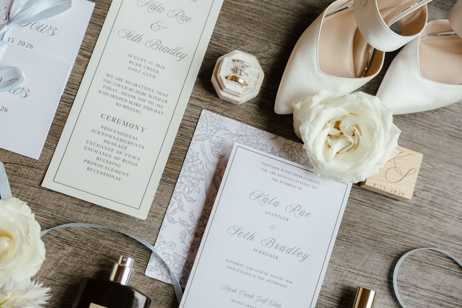 Elegant wedding flat lay with floral invitations, white rose, perfume bottles, rings, and bridal heels.