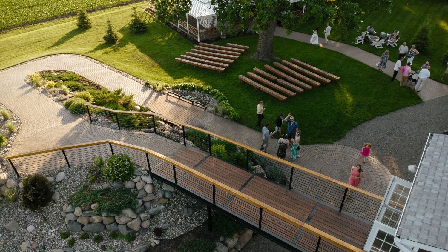 Aerial view of a garden wedding venue with wooden bridge, amphitheater seating, and guests on a summer evening.