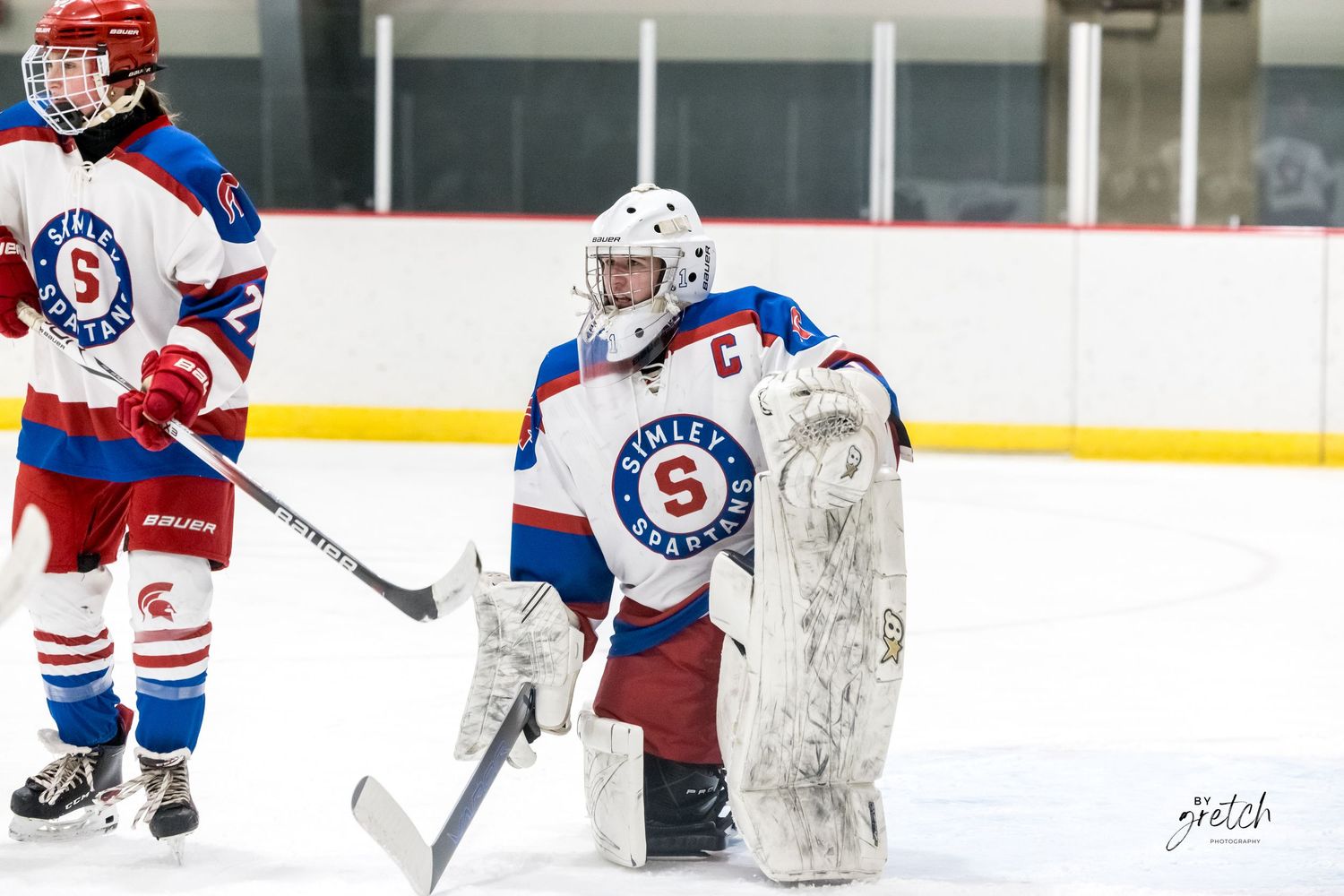 Youth hockey goalie in red white and blue uniform with captain C patch on ice.