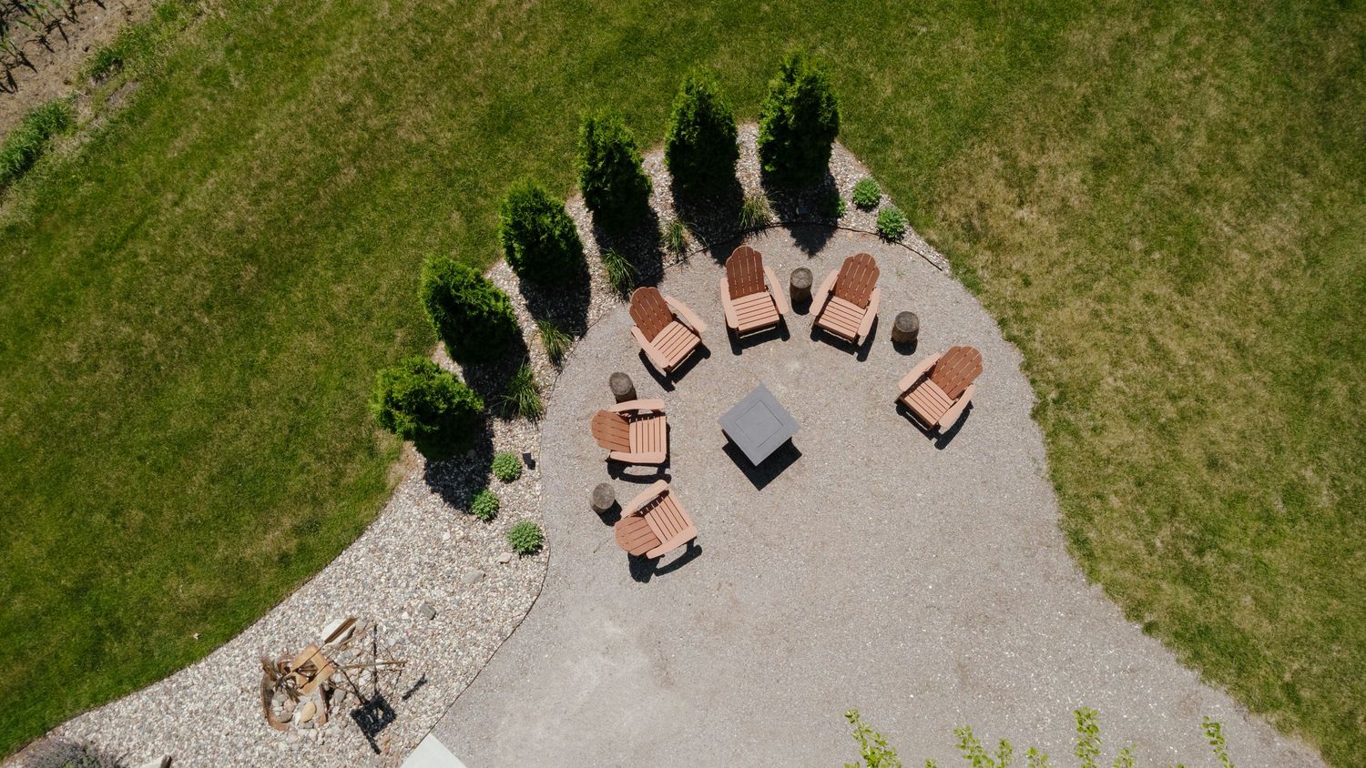 Aerial view of outdoor seating area with wooden chairs arranged around a fire pit on gravel, surrounded by green lawn.
