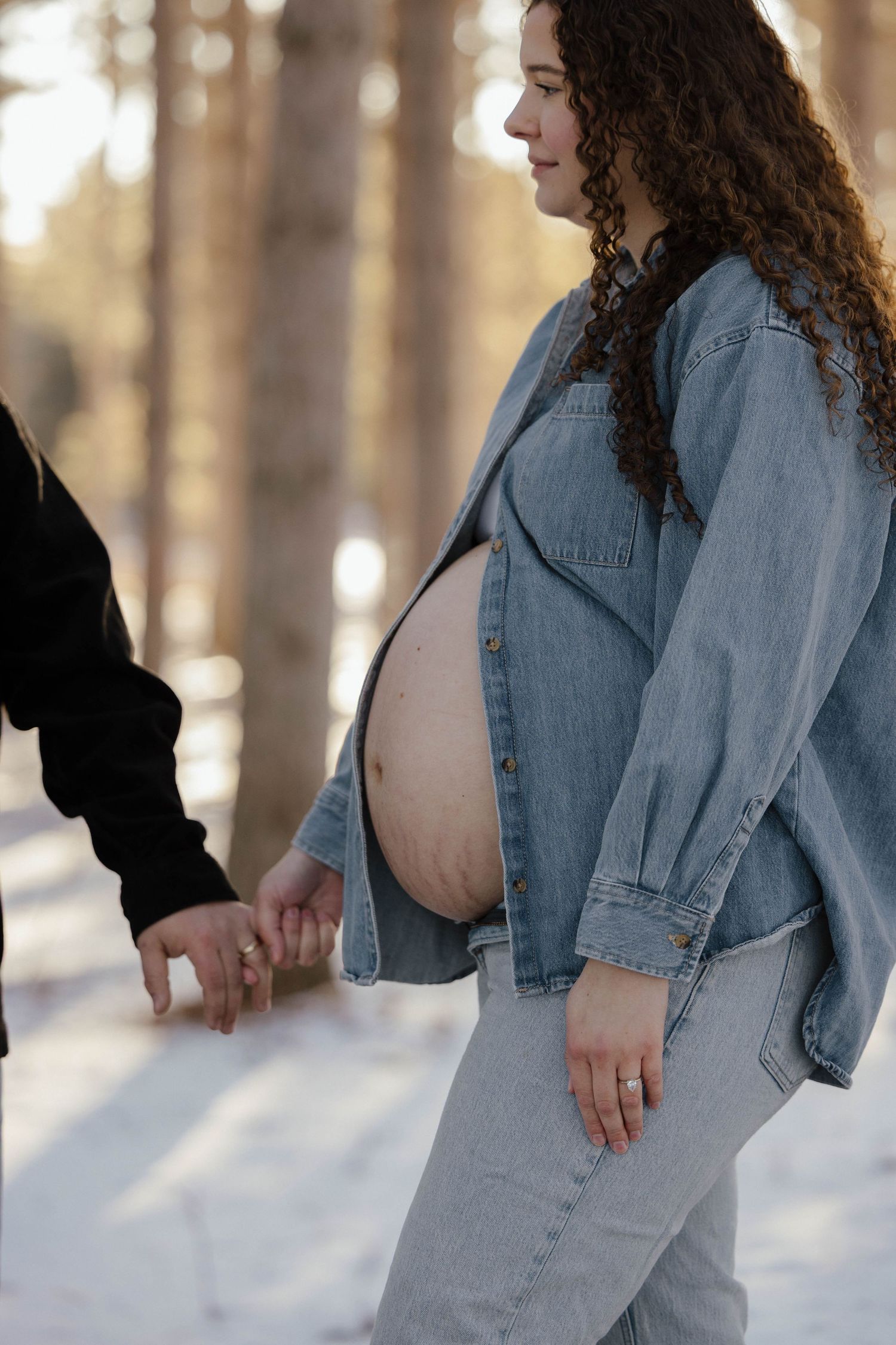 Pregnant woman in denim shirt and jeans holding hands with partner while showing bare baby bump in snowy forest.