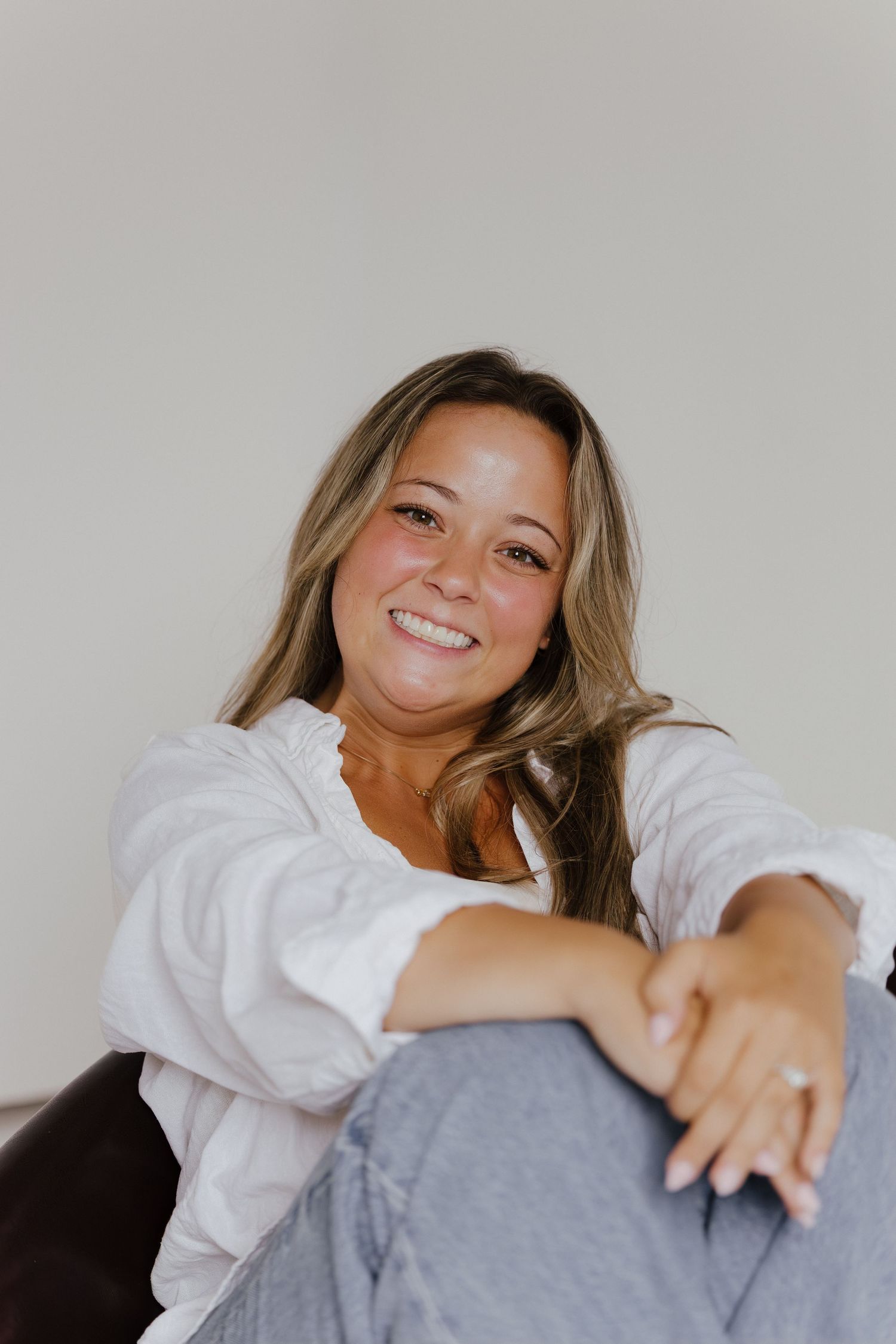 Smiling woman in white shirt and gray jeans sitting relaxed on floor.