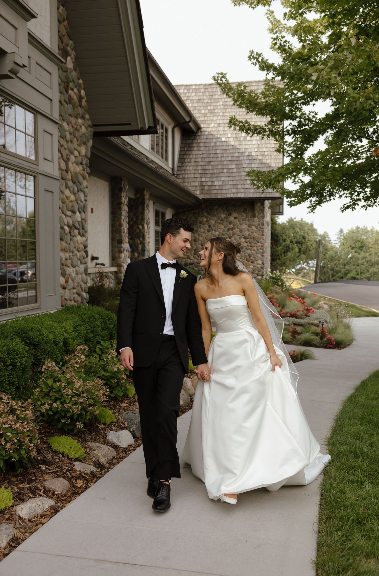 Bride and groom walking hand in hand along a path beside a stone building, bride in strapless white gown.