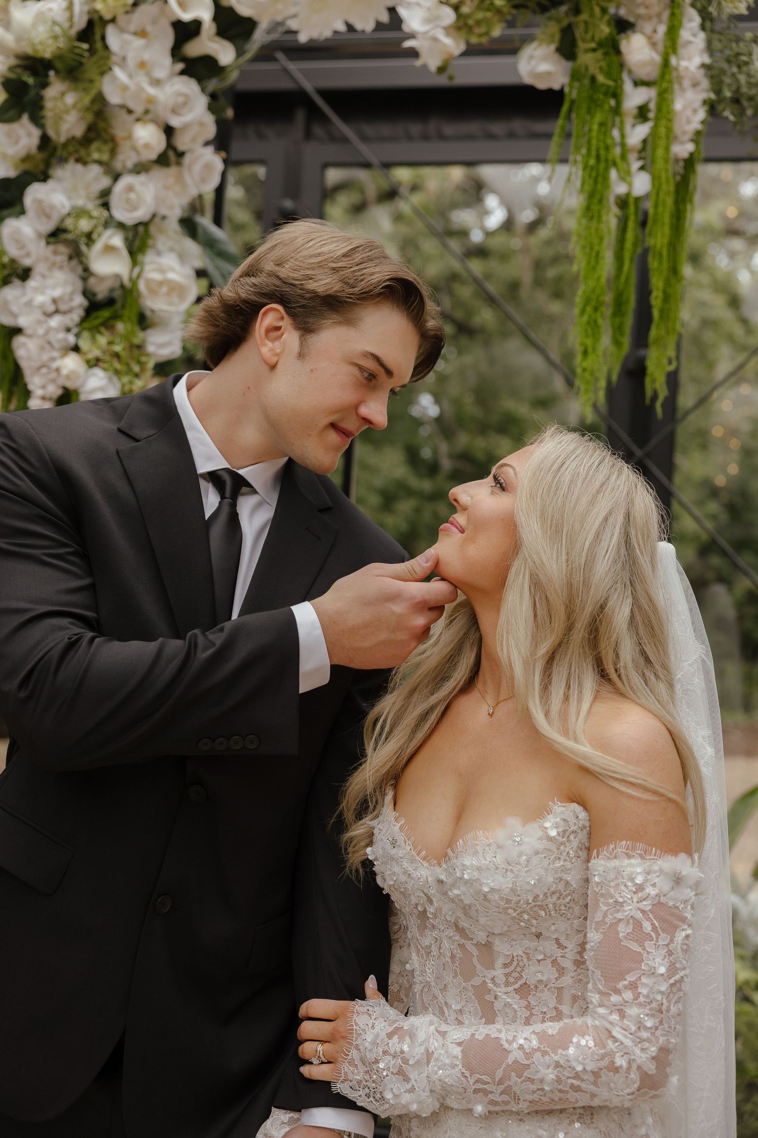 Groom tenderly touching bride's face under a white floral arch during their outdoor wedding ceremony.
