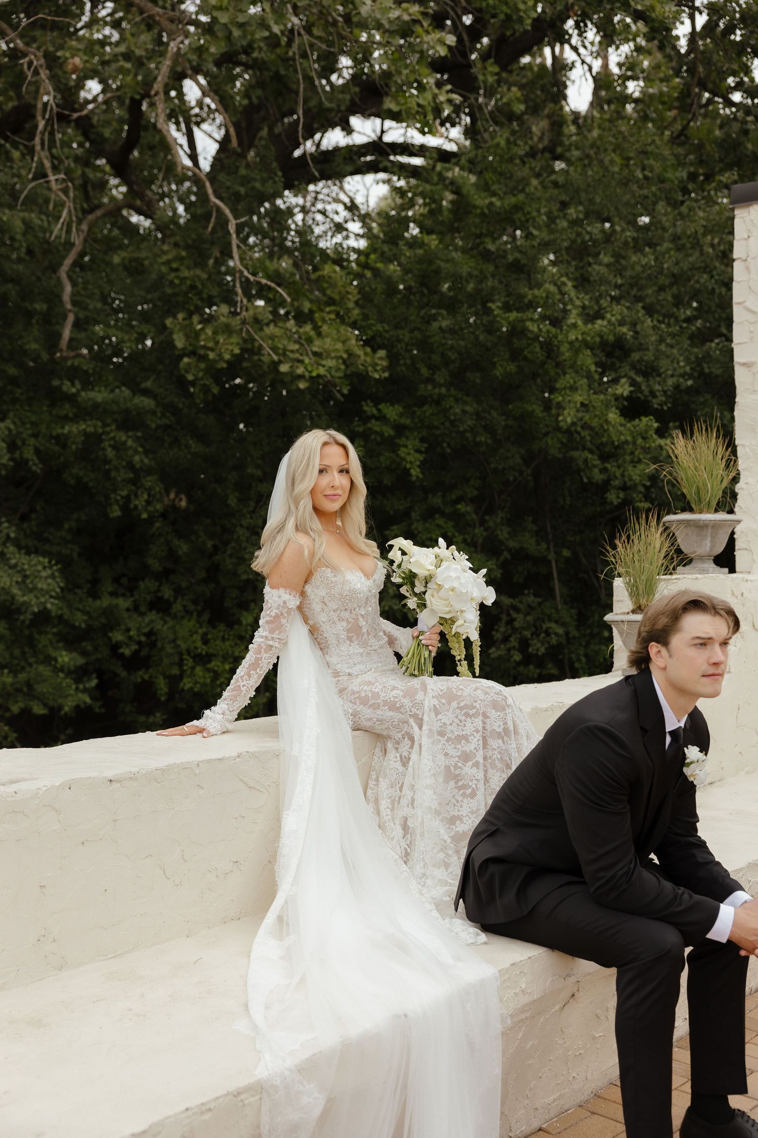 Bride in long-sleeve lace gown holding white bouquet poses with groom on white stone outdoor terrace.