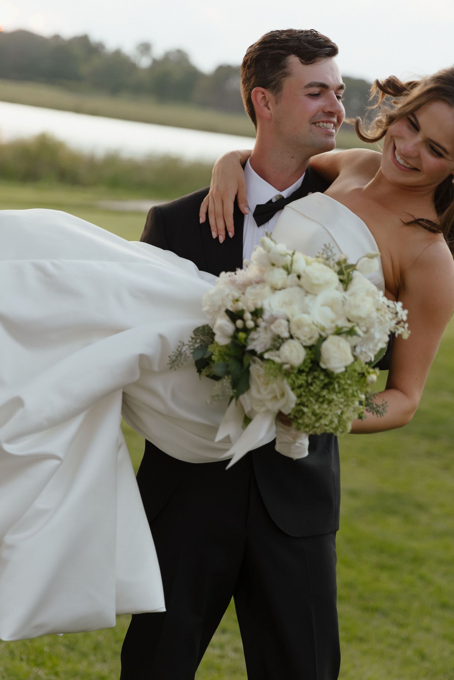Groom carrying laughing bride in white gown and bouquet of white roses across a green lawn near a lake.