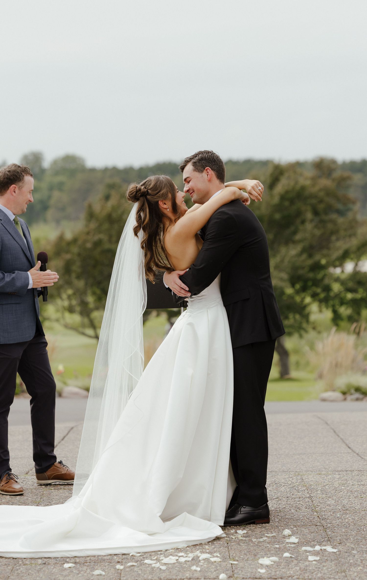 Bride and groom share first kiss at outdoor ceremony while officiant stands nearby smiling.