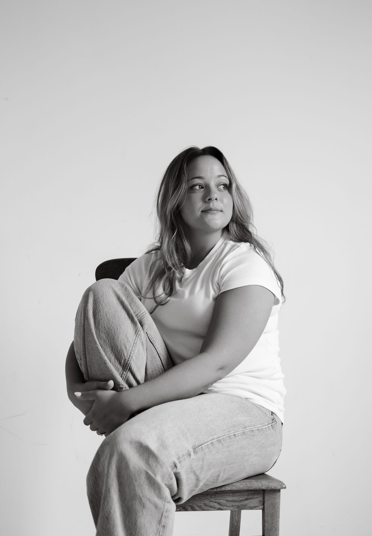Black and white portrait of young woman in white tee and jeans sitting on stool with knees drawn up.