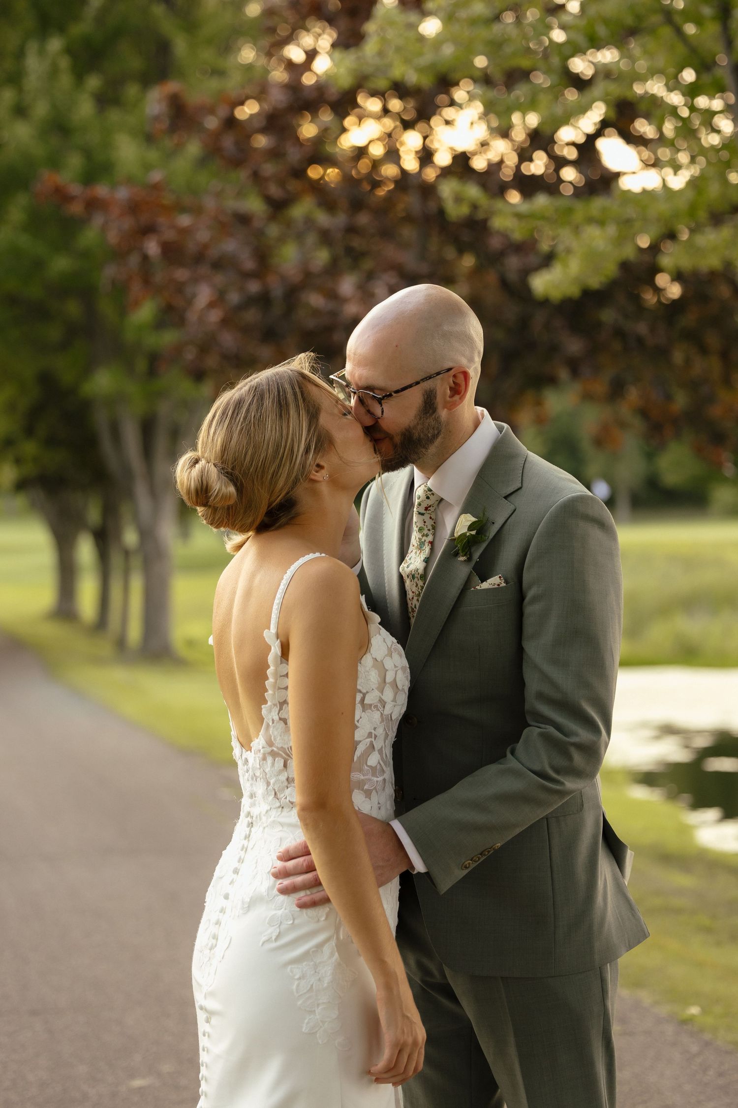 Bride and bald groom share romantic kiss on tree-lined path in golden evening light at outdoor wedding.