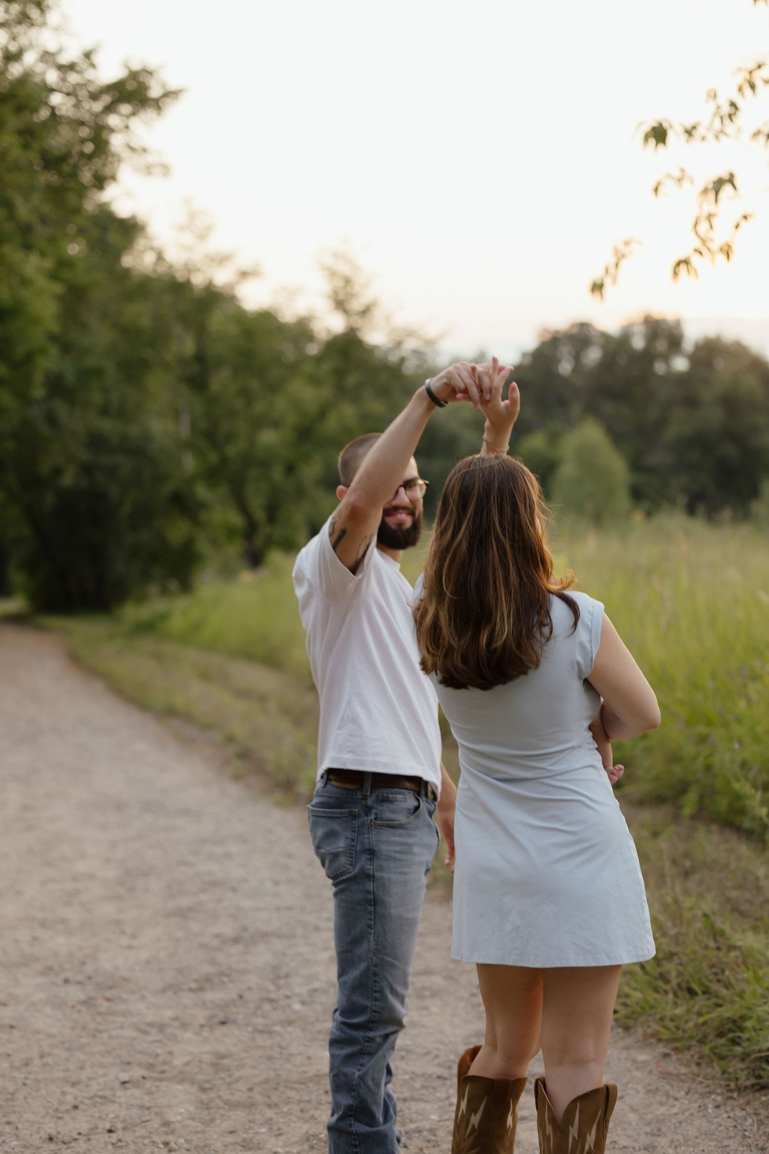 Couple dancing on a dirt path surrounded by lush greenery, man in white tee and jeans, woman in white dress and brown boots.