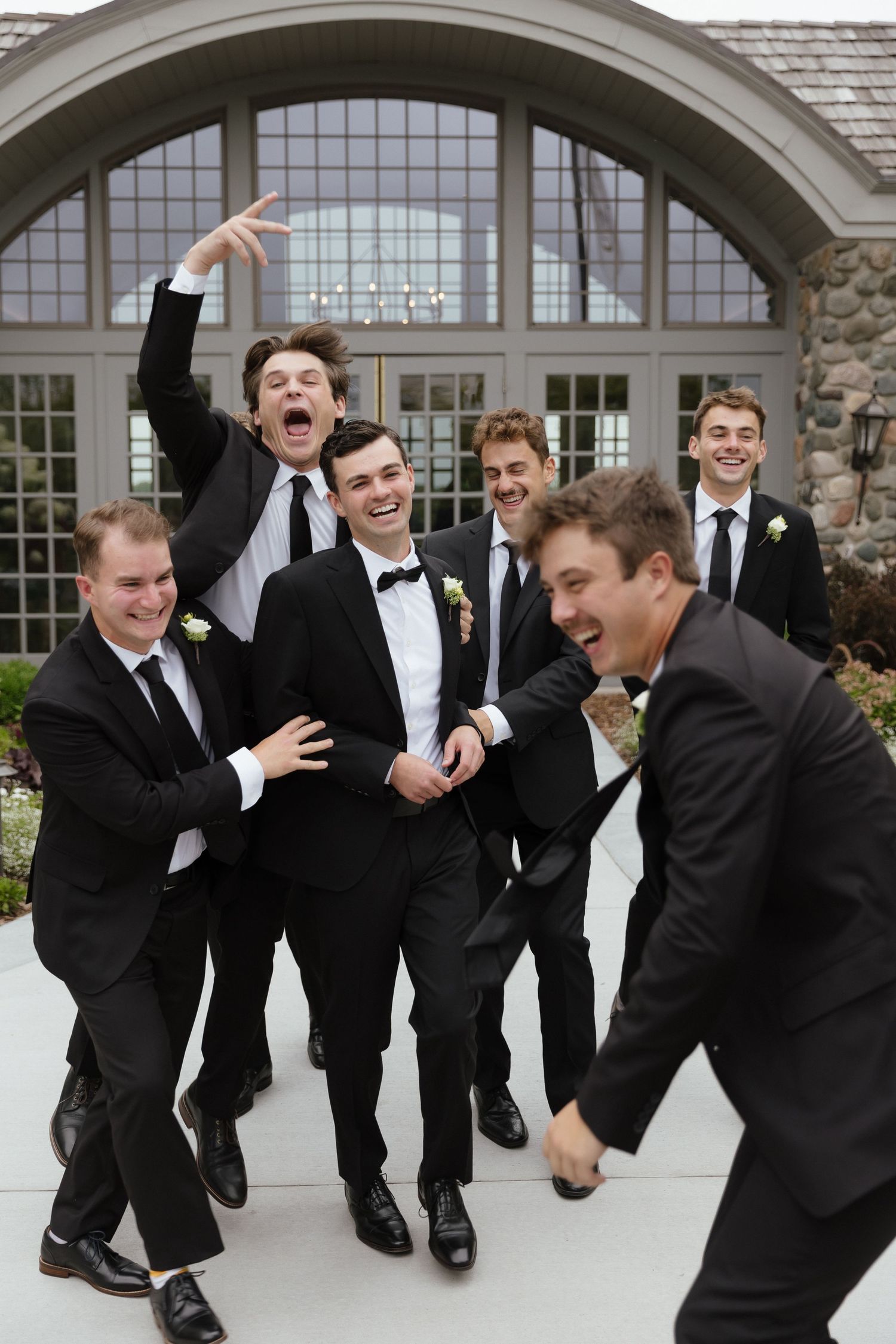 Groom laughs joyfully with energetic groomsmen in black tuxedos outside stone wedding venue entrance.