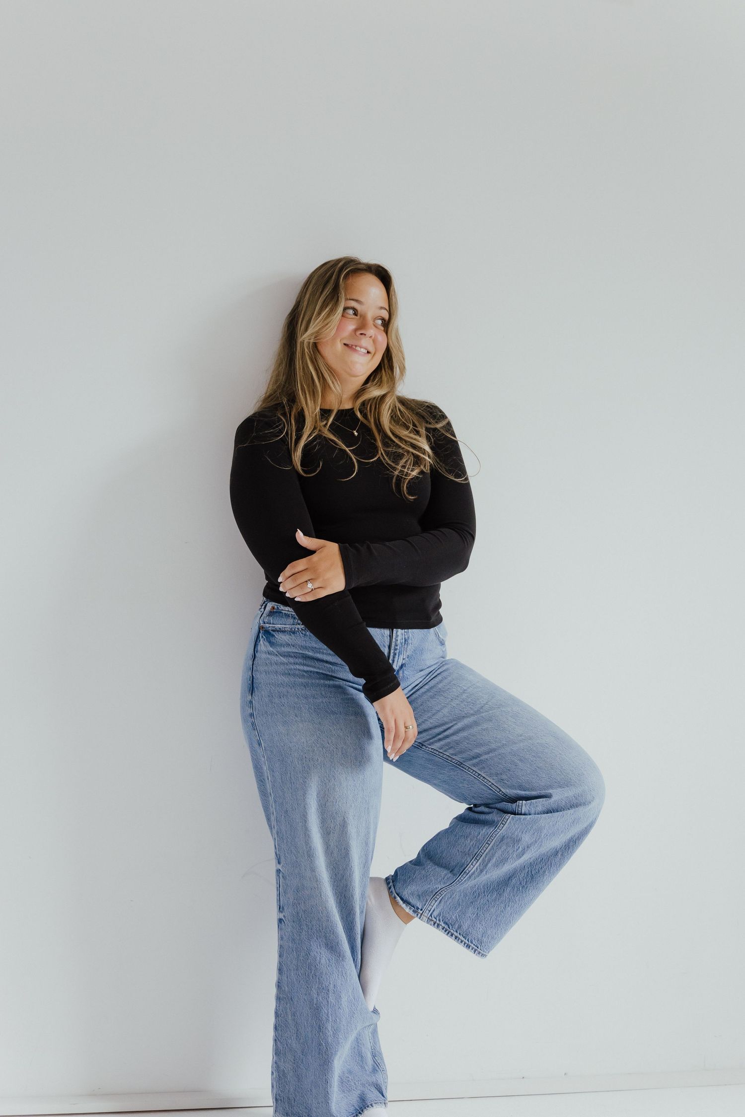 Woman in black sweater and wide-leg jeans posing against white wall smiling.
