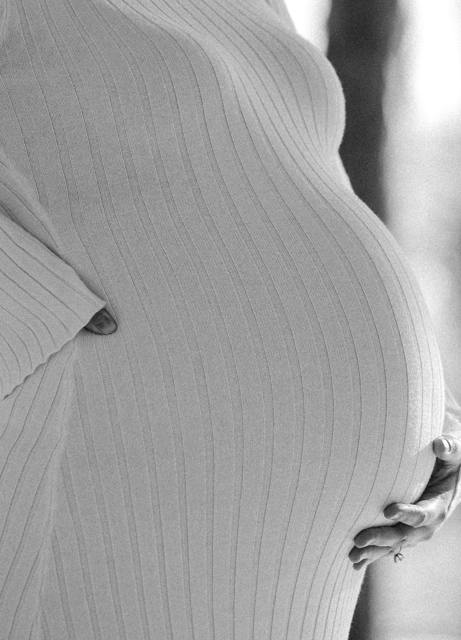 Close-up black and white photo of a pregnant woman in a ribbed white dress cradling her baby bump.