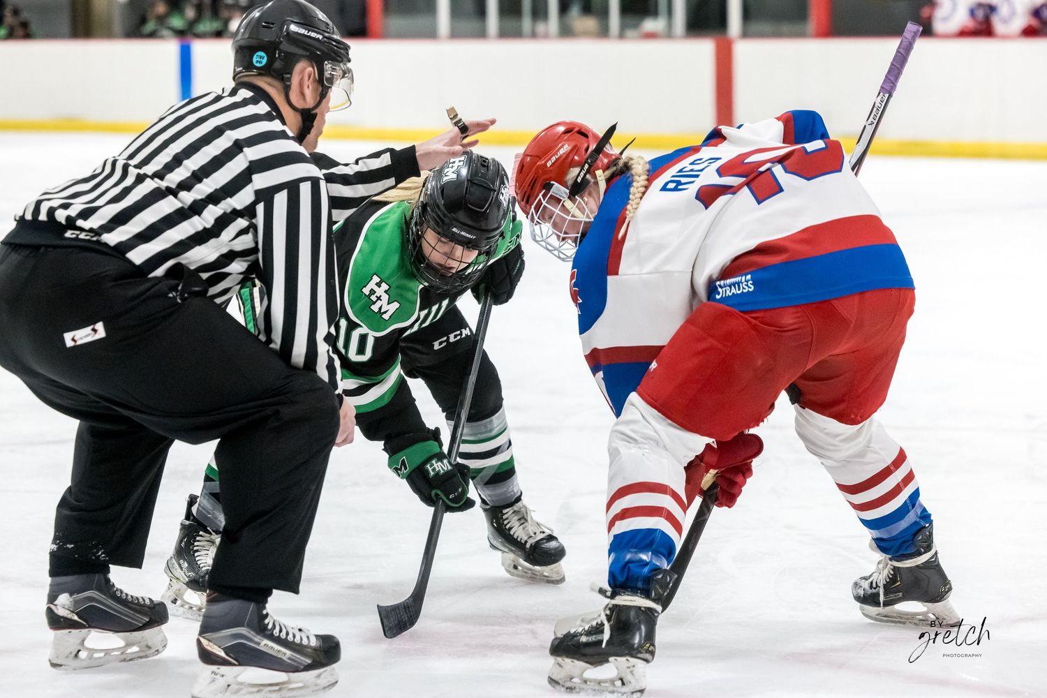 Hockey referee oversees a face-off between green and red-white-blue uniformed players.