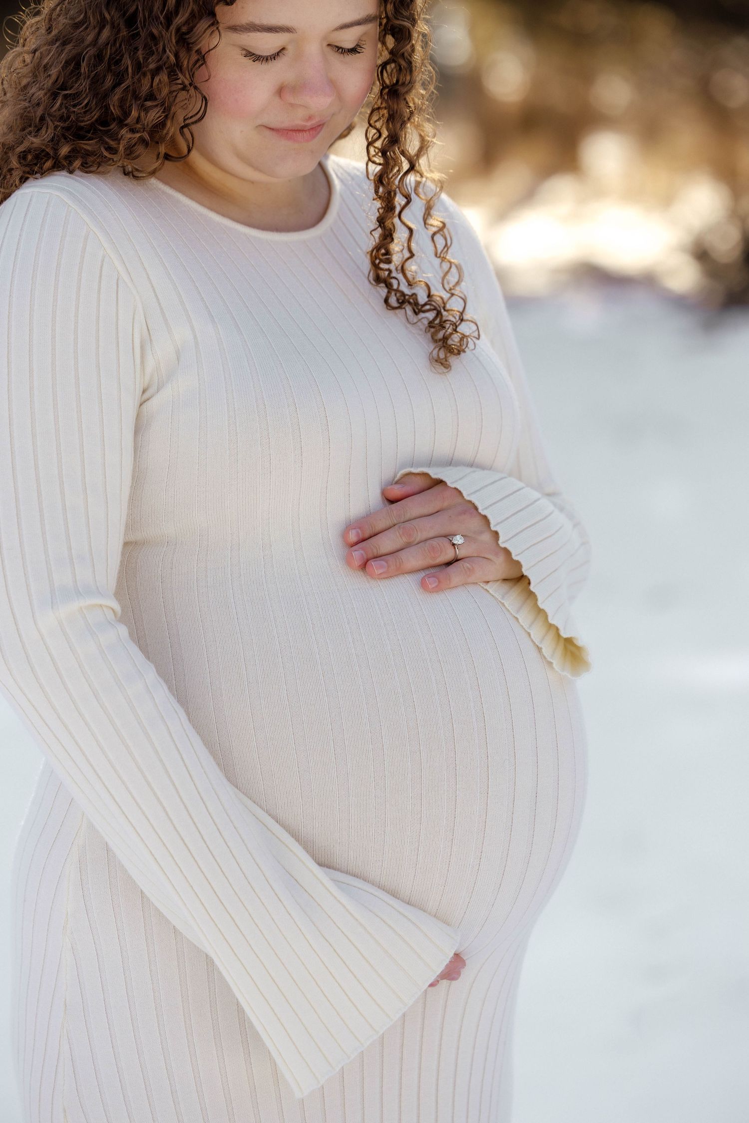 Pregnant woman with curly hair in a white ribbed dress holding a baby hat against her bump outdoors in snow.