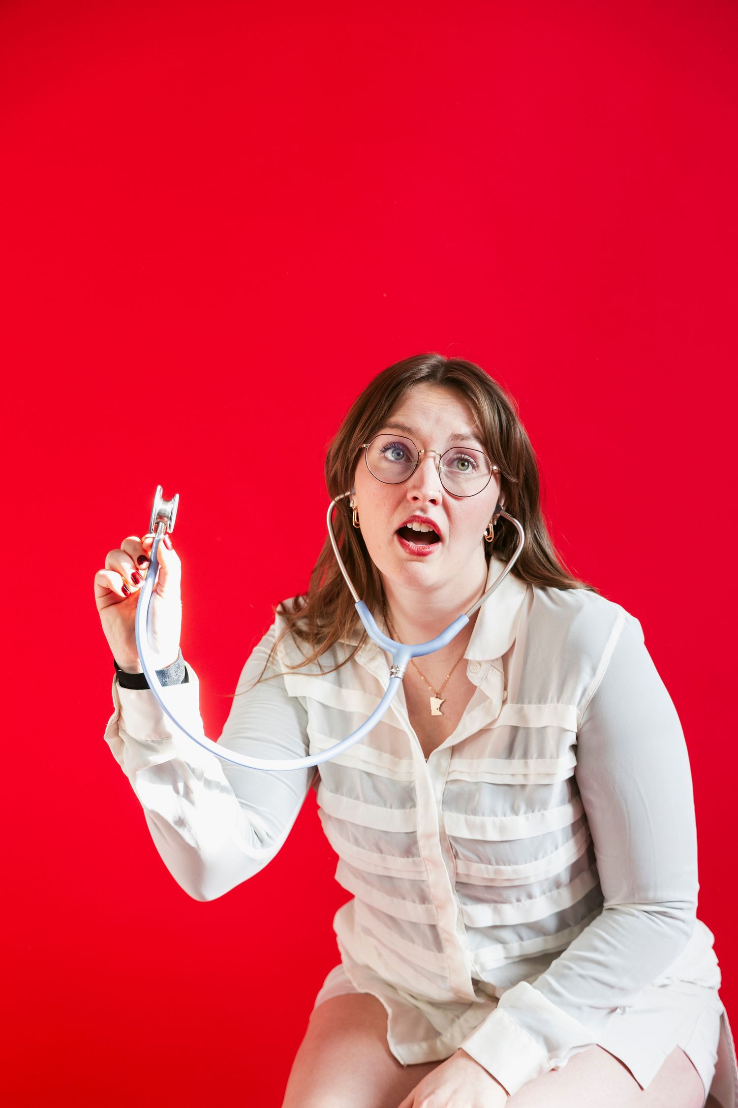 Woman in white dress wearing glasses holding a stethoscope with surprised expression against red background.