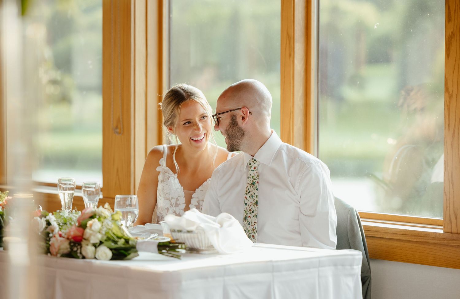 Smiling bride and groom seated together at sweetheart table during wedding reception.