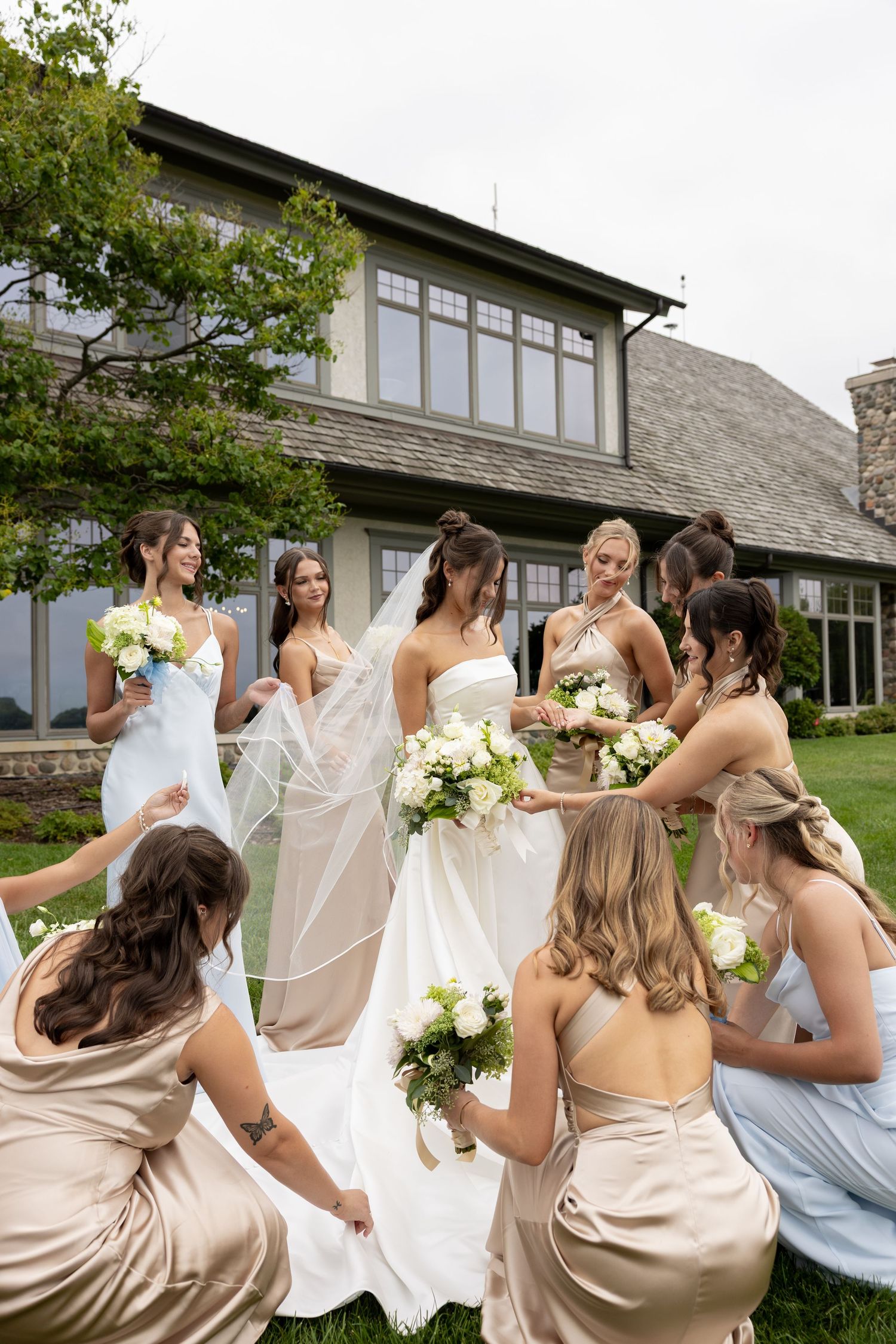 Bride surrounded by bridesmaids in champagne and blue gowns holding white bouquets on green lawn outside elegant venue.