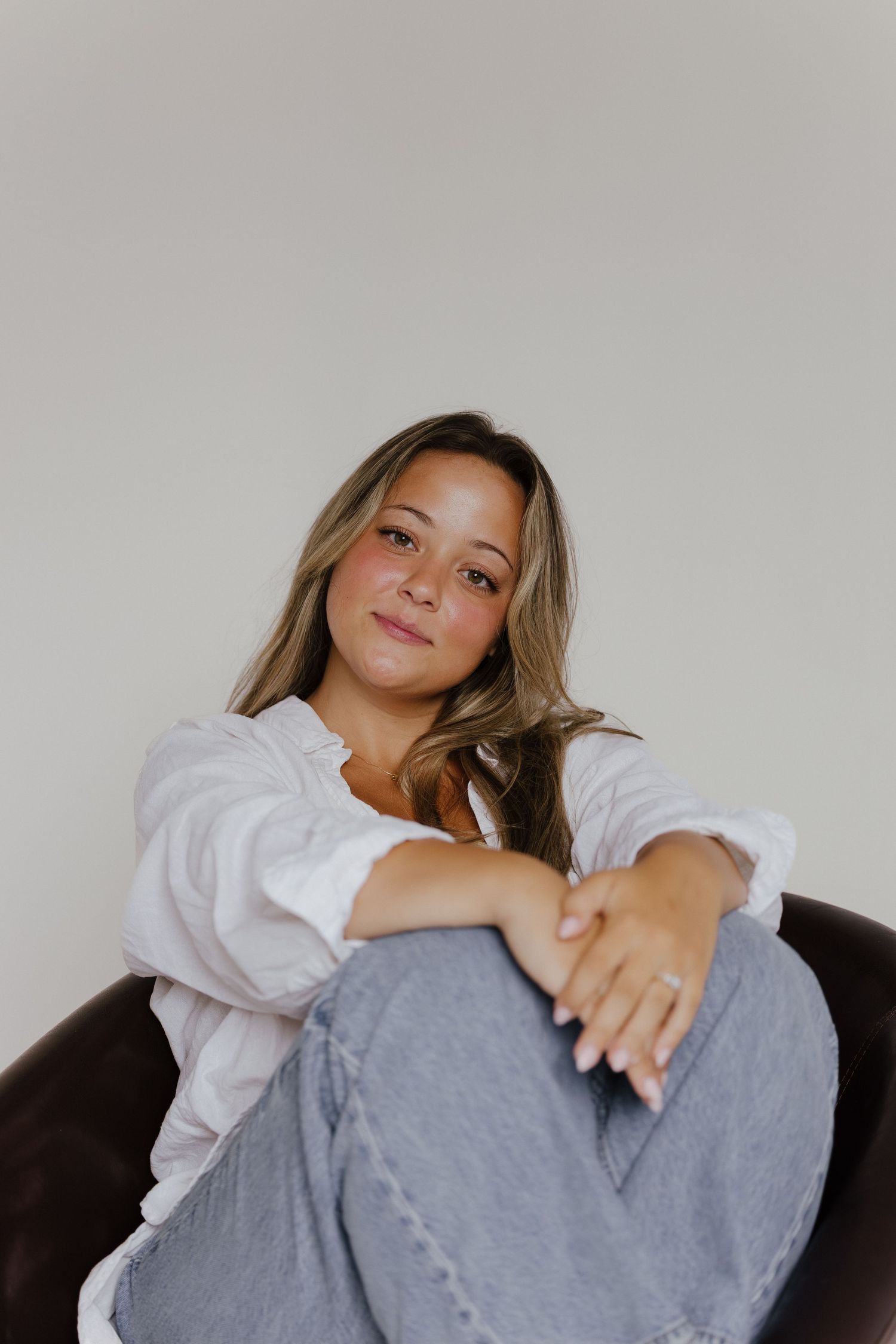 A young woman in a white shirt and jeans sits relaxed in a dark chair against a neutral background.