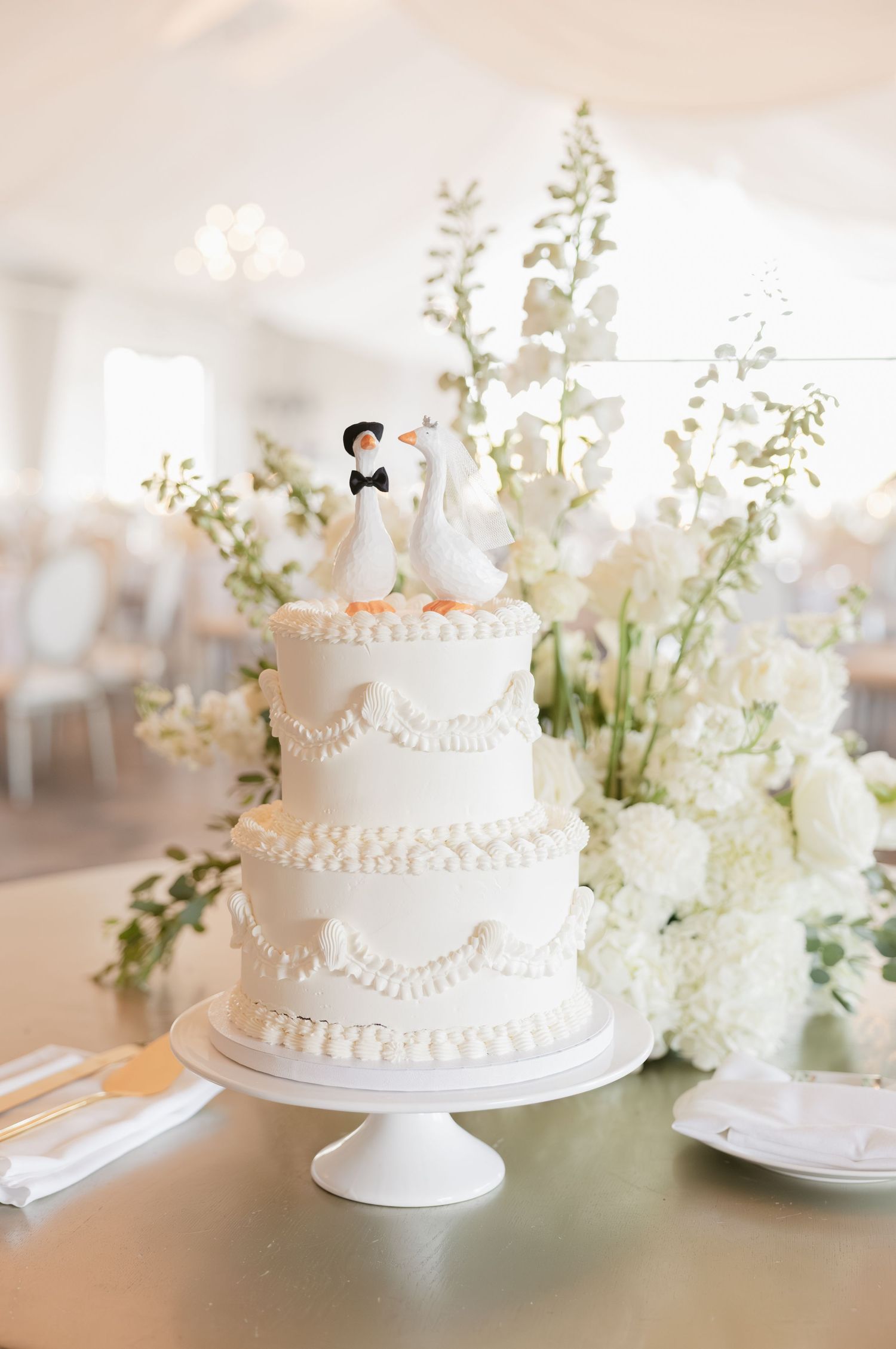 Elegant three-tier white wedding cake with pearl details and duck bride and groom cake toppers surrounded by white floral arrangements.