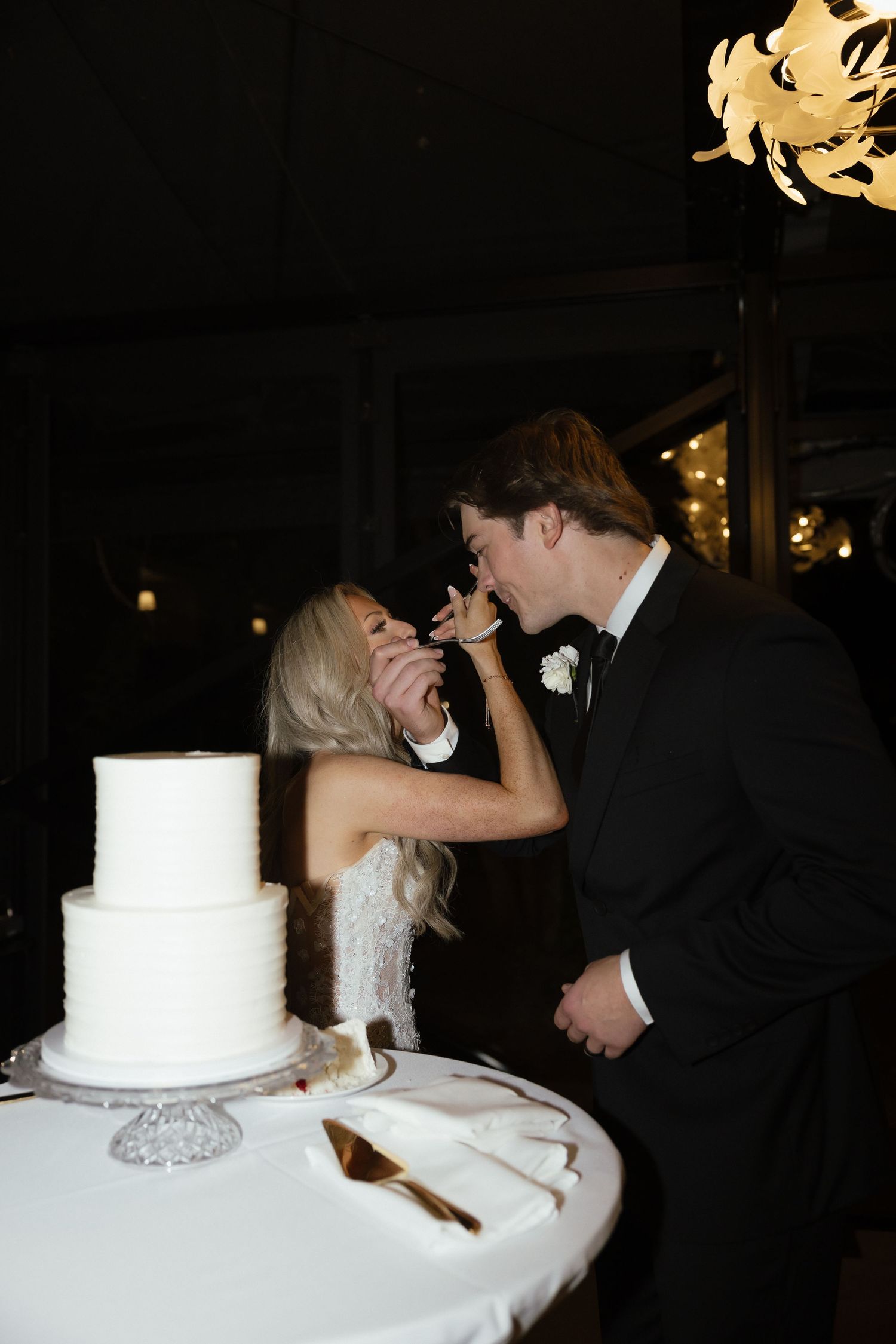 Bride and groom sharing wedding cake at reception beside a two-tier white frosted cake.