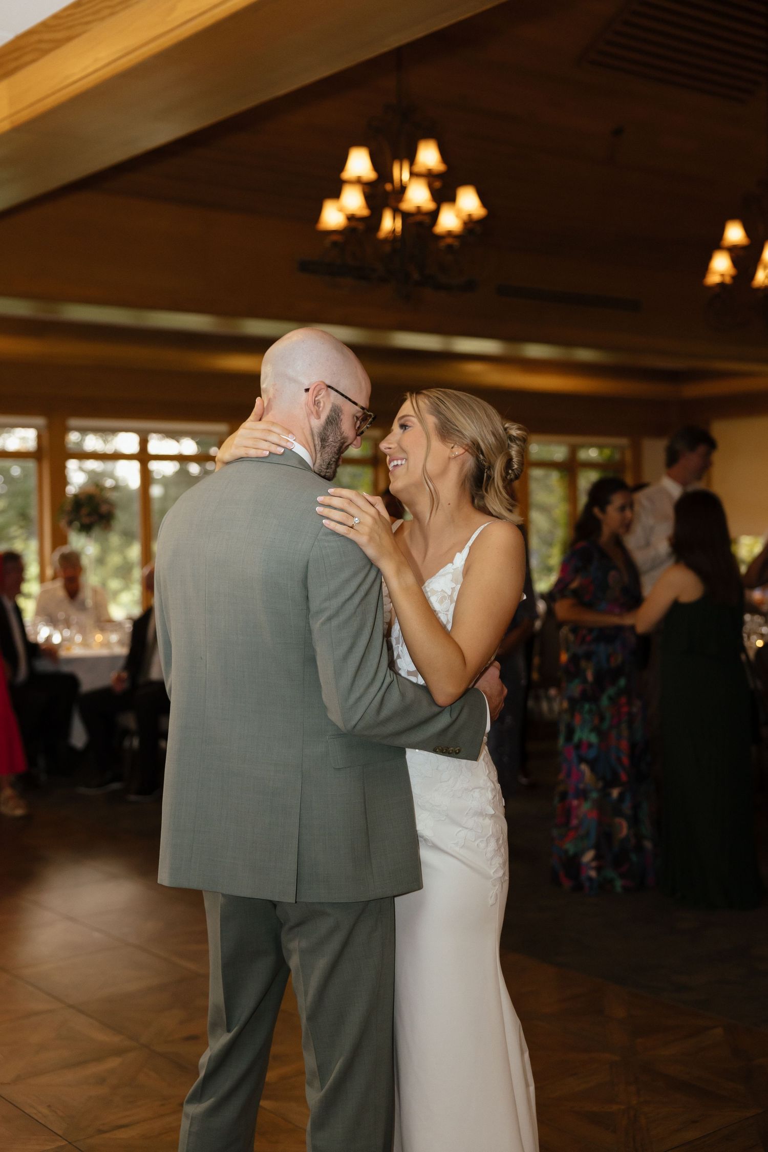 Bride in lace gown smiles while sharing first dance with groom in gray suit inside warmly lit reception hall.