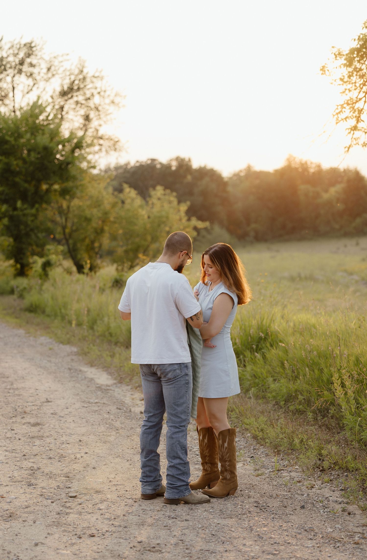 Couple standing on a dirt path at golden hour, woman in white dress and cowboy boots, man in white shirt and jeans.