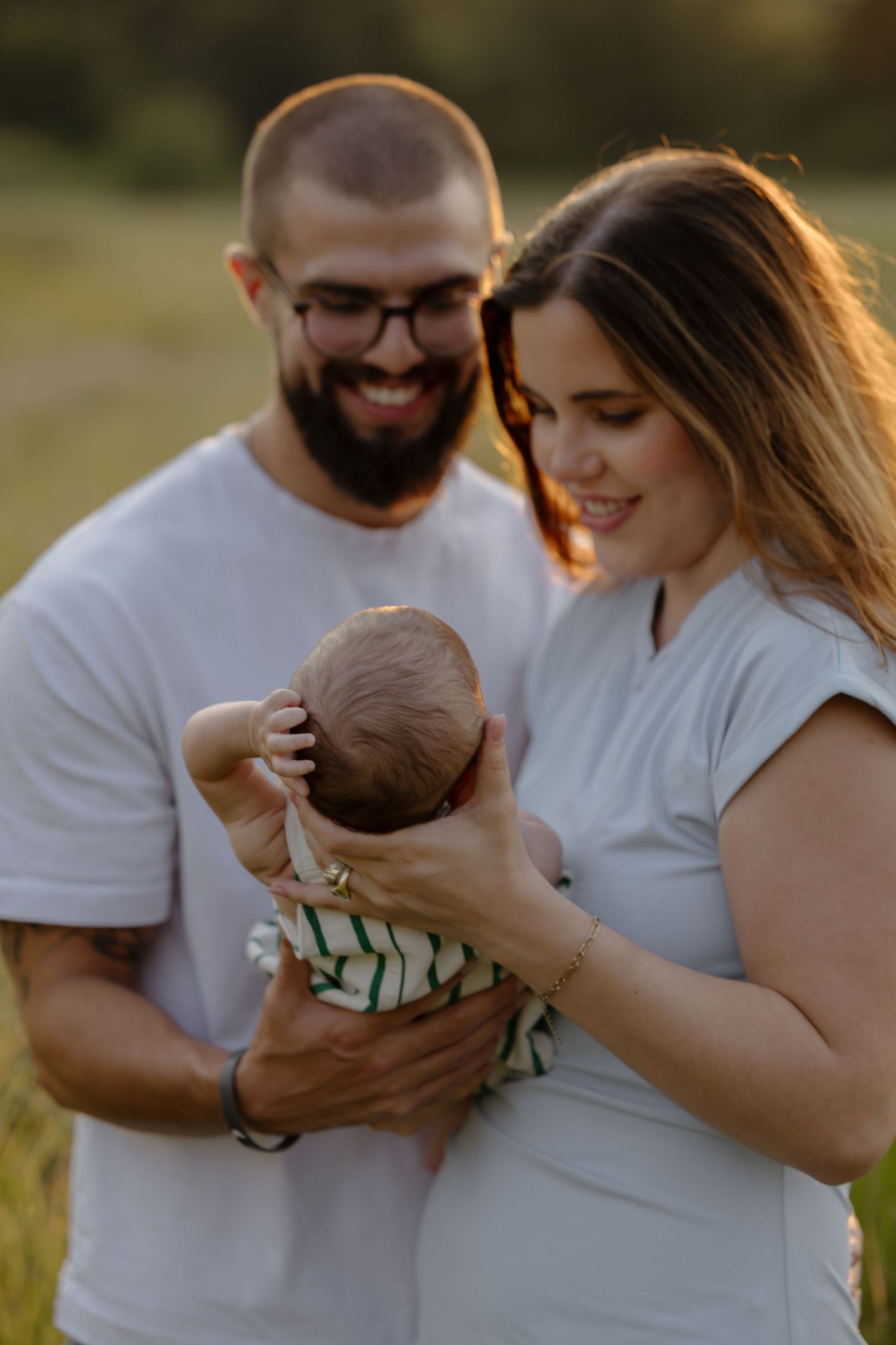 Smiling couple holding newborn baby outdoors in golden hour light, father in white tee and glasses, mother in light blue dress.