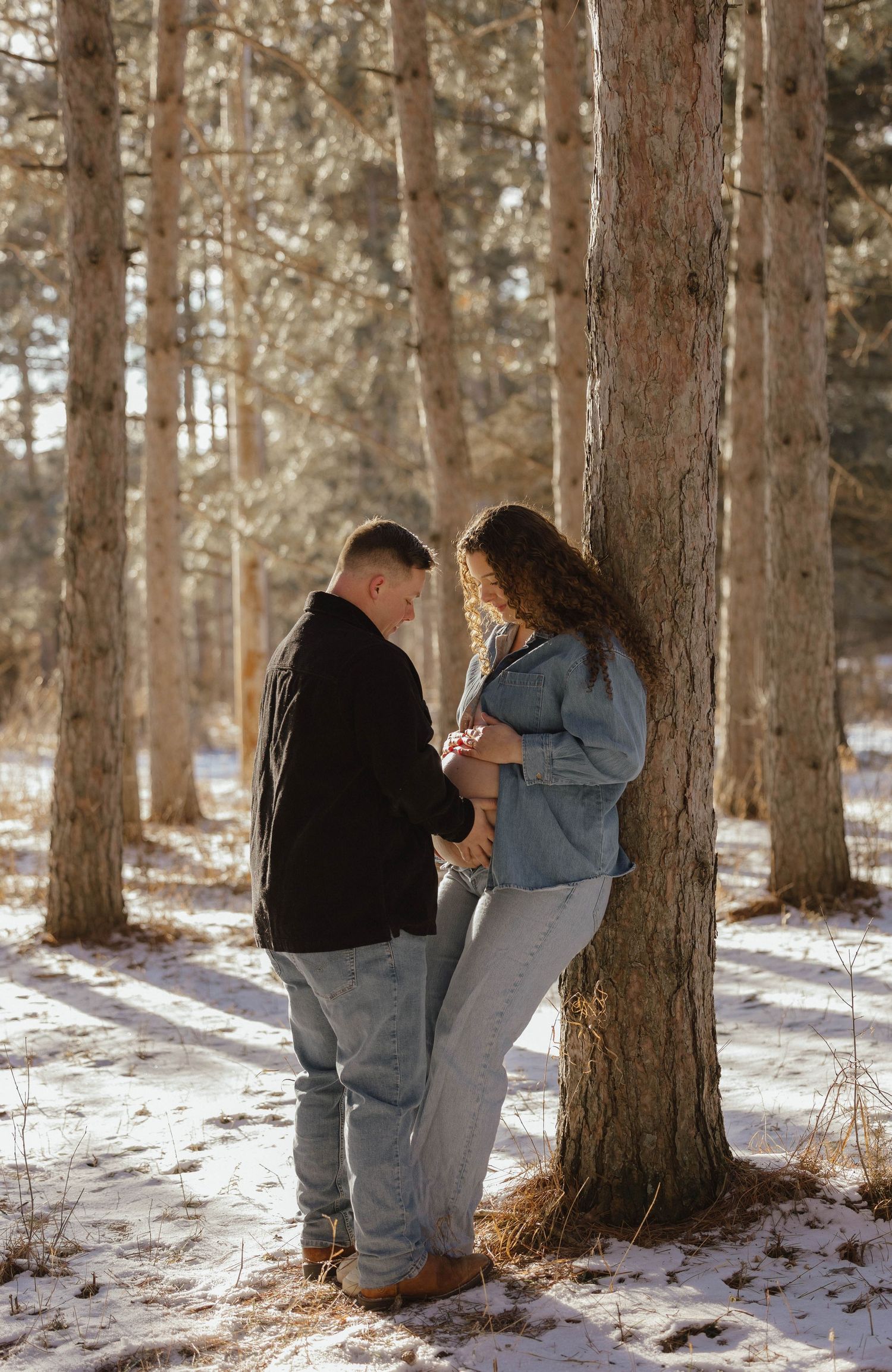 Expecting couple standing together against a tree in a snow-covered forest during a winter maternity photoshoot.