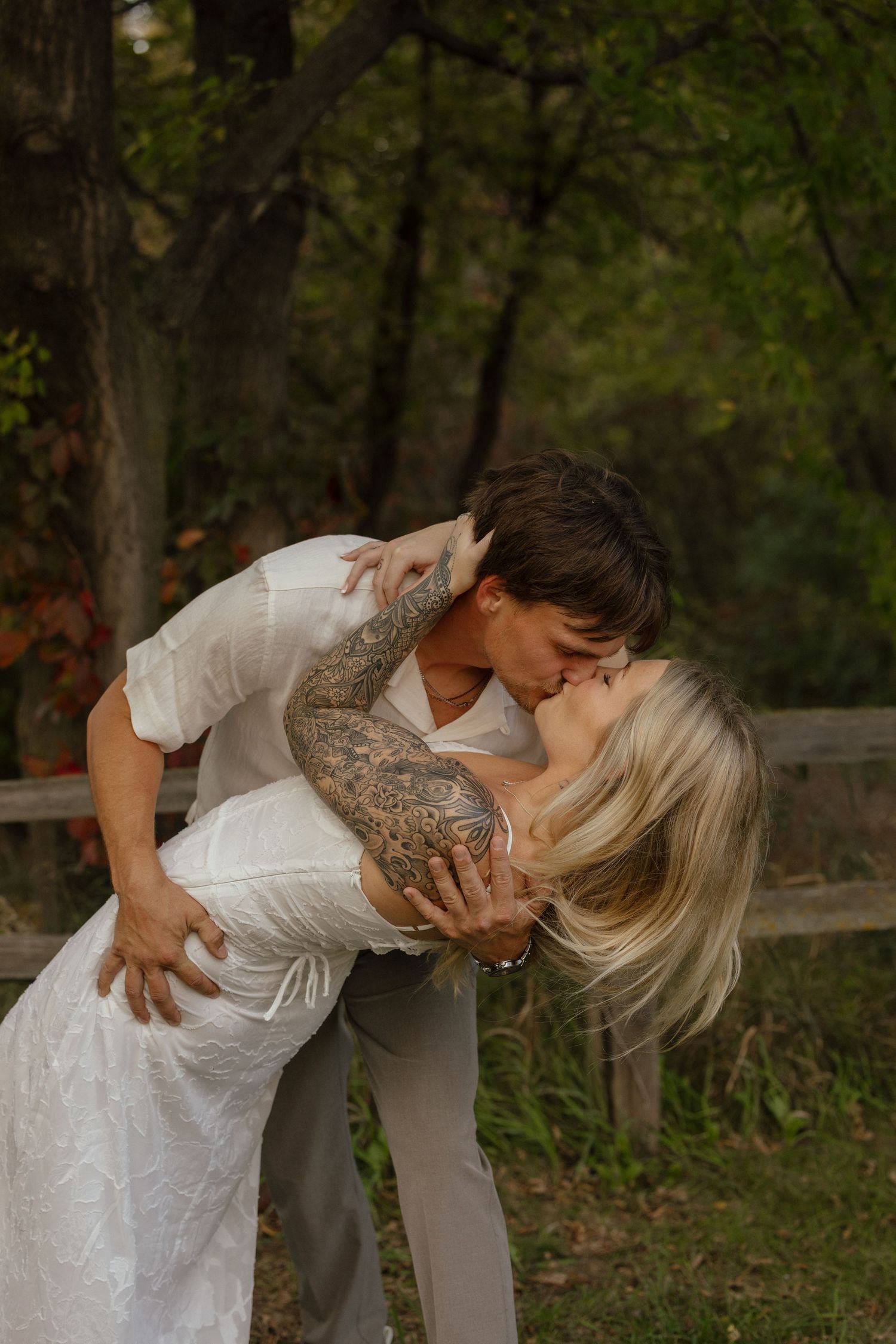 Groom with tattoos dipping and kissing blonde bride outdoors among autumn trees.