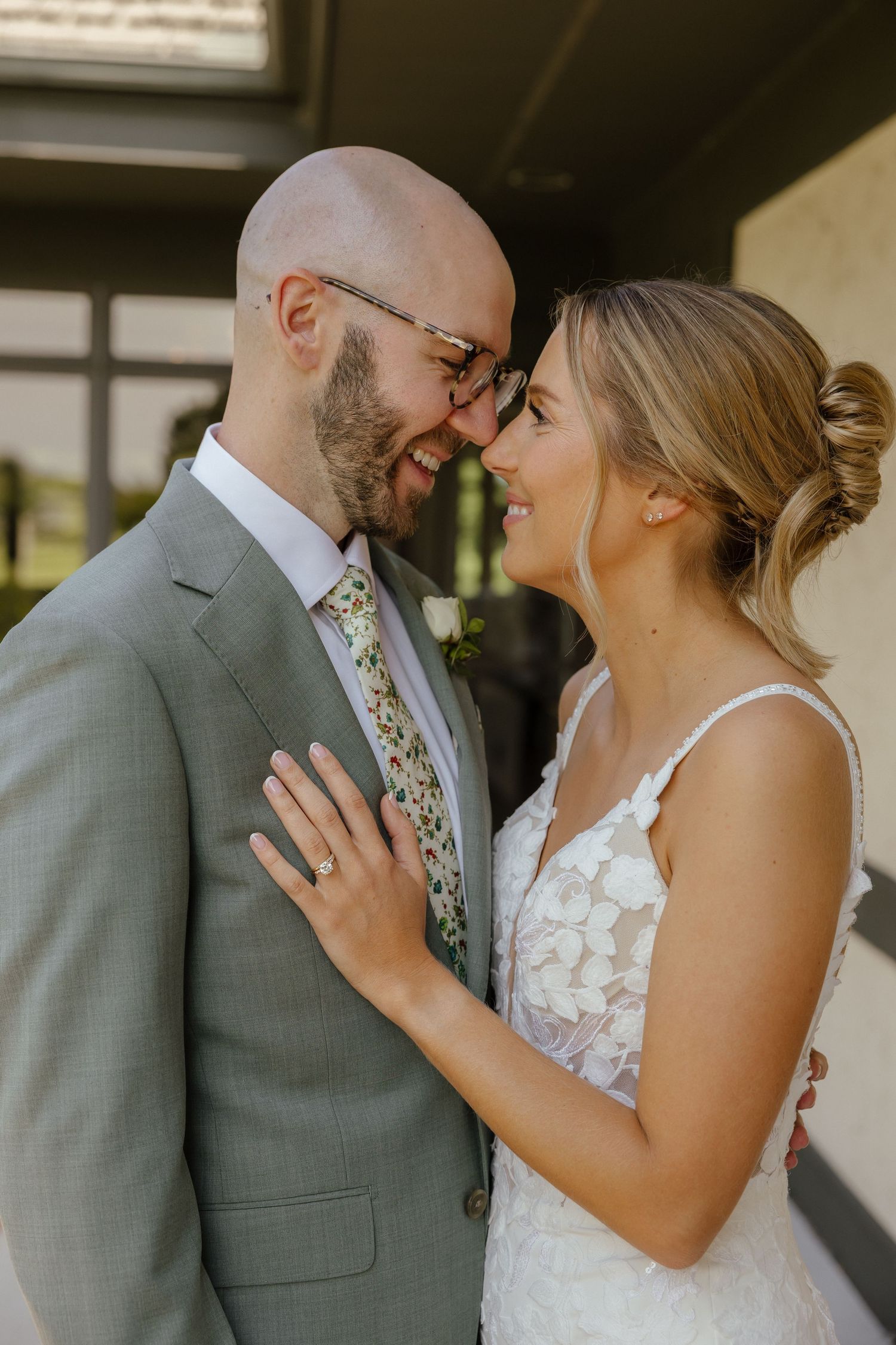 Bride in lace gown and groom in gray suit sharing intimate moment touching foreheads outdoors.