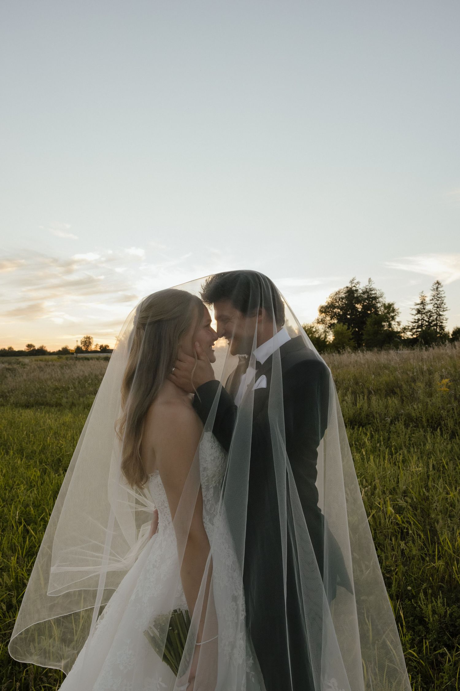 Bride and groom sharing an intimate moment under a flowing veil in a golden sunset meadow.