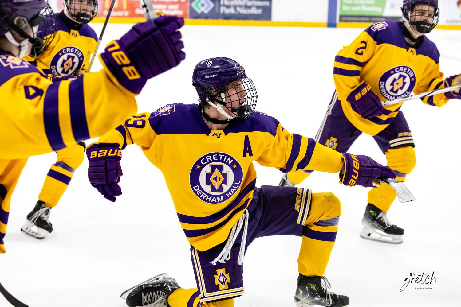 Hockey player in purple and gold celebrating goal on knees on ice rink.