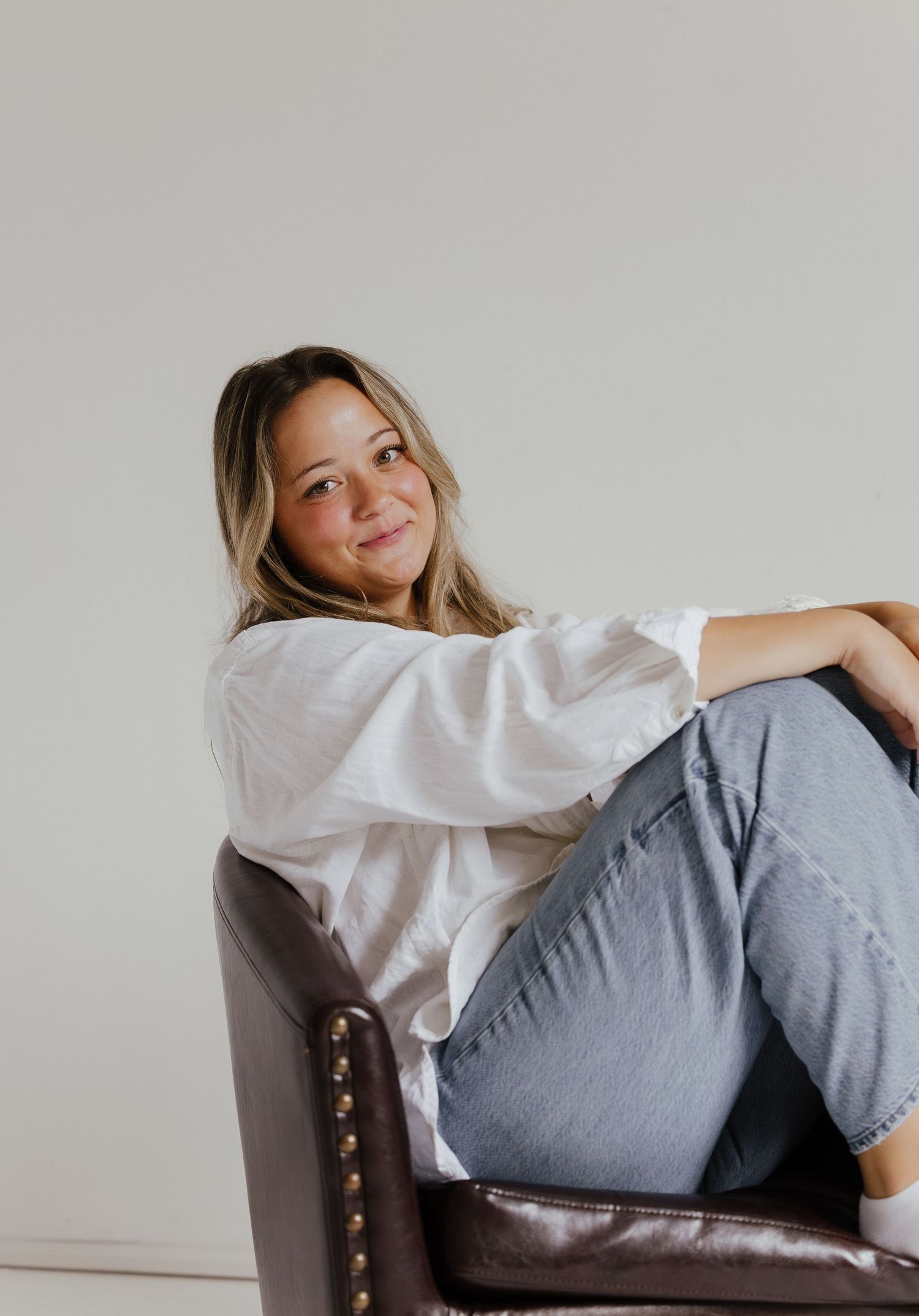 Young woman in white button-up shirt and jeans relaxing in brown leather chair, smiling softly.