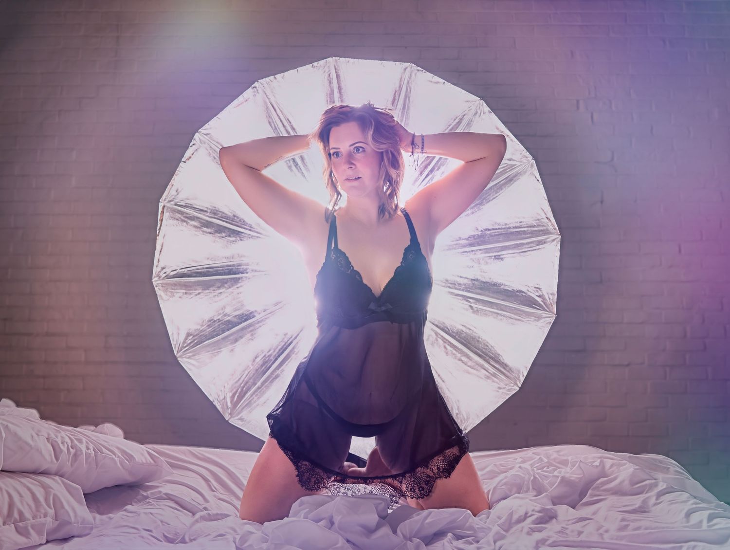 Woman in black lace lingerie kneeling on bed posed in front of large round reflective photography light.